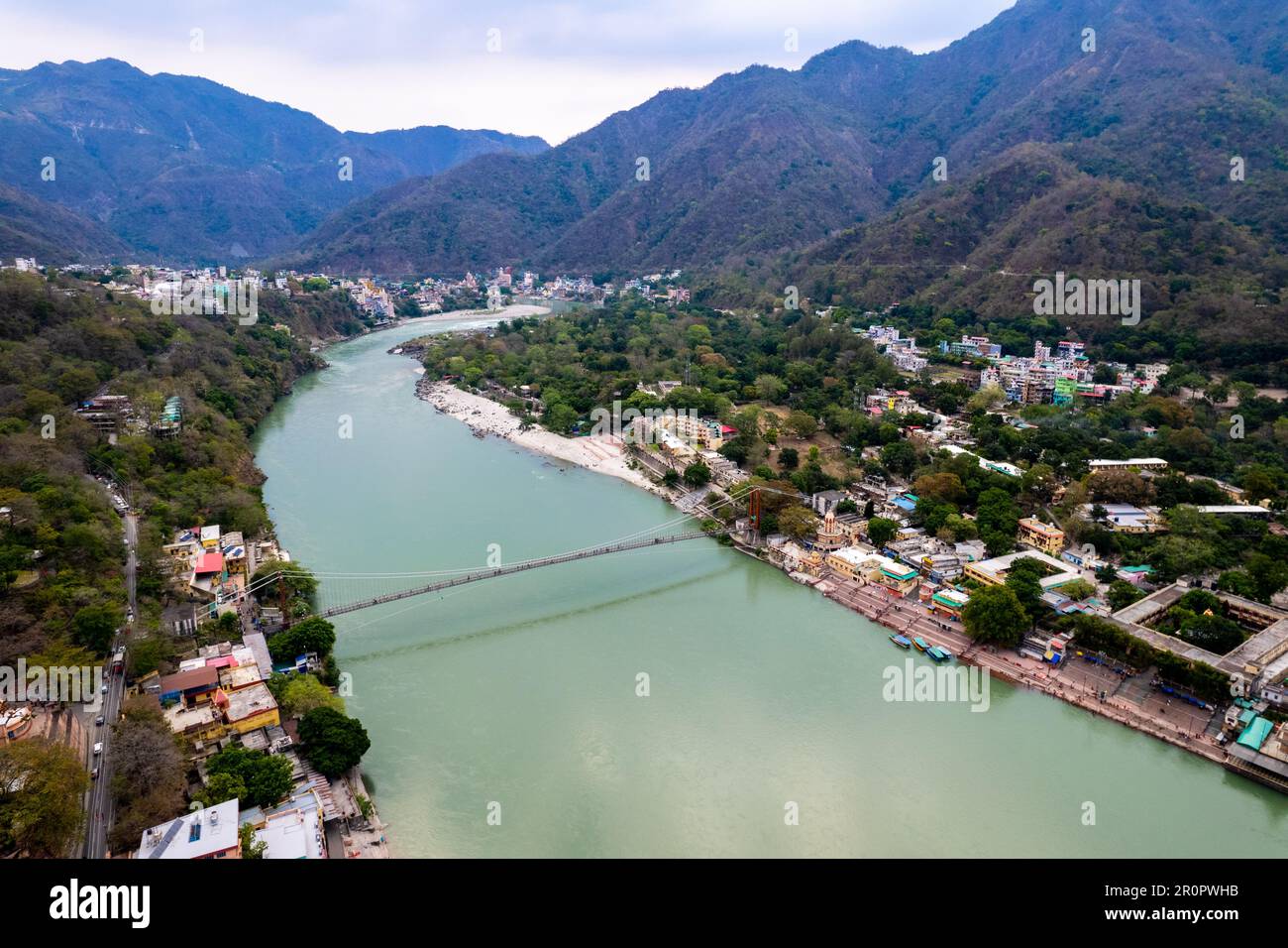 aerial drone shot of blue water of river ganga stretching into distance with himalayas with ram setu suspension bridge and temples on the banks of the Stock Photo