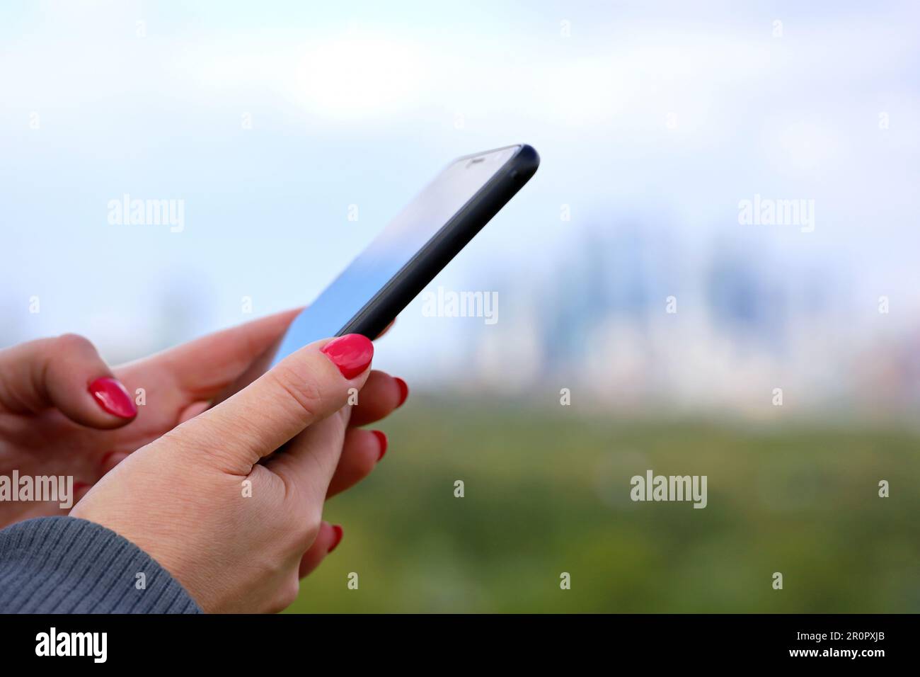 Female hands with smartphone close up on blurred city skyscrapers background. Woman with red manicure using mobile phone in park Stock Photo