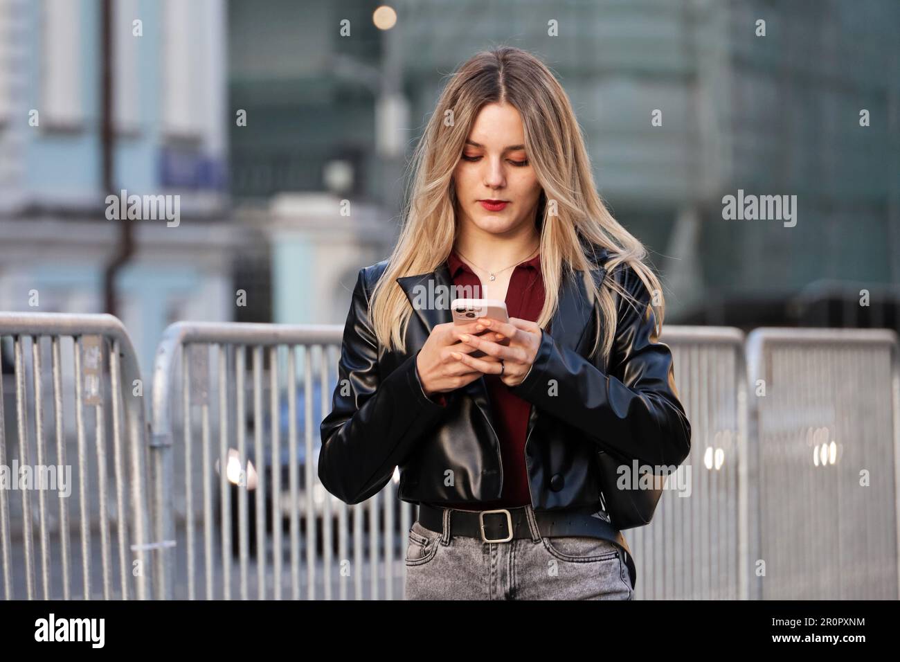 Blonde girl in black jacket walking down a street with smartphone. Using mobile phone in spring city Stock Photo