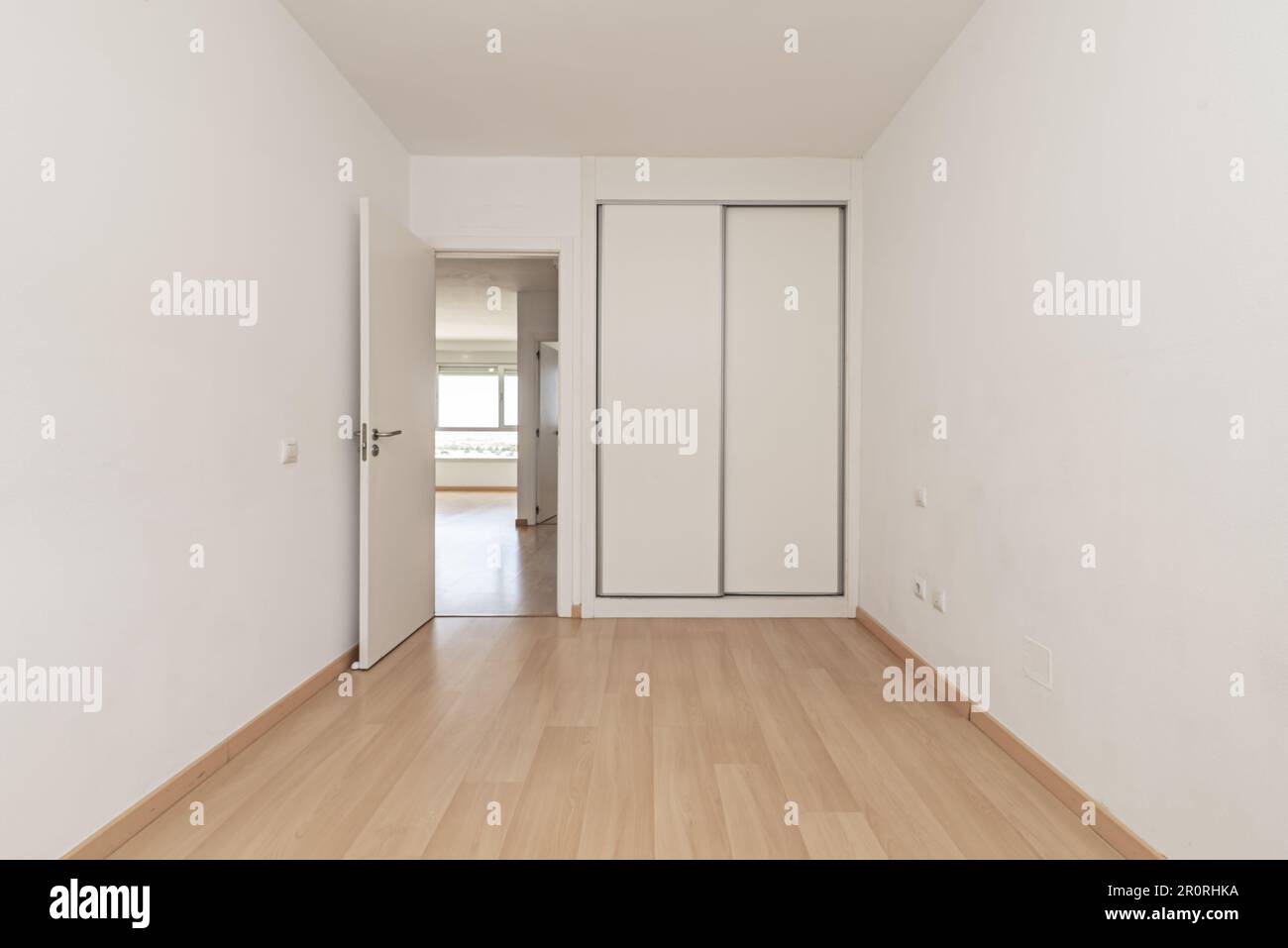 An empty room in an apartment with a built-in wardrobe with white sliding doors, a light wooden floor and white access doors, plain walls and wooden b Stock Photo