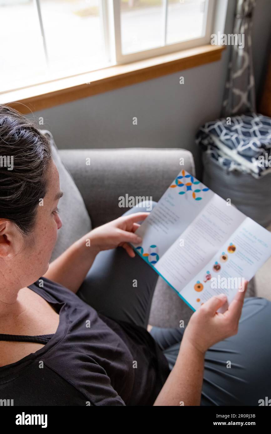 Top down view of 30s female reads skin cancer literature from dermatology department on couch at home upon recent skin cancer diagnosis on her chest. Stock Photo