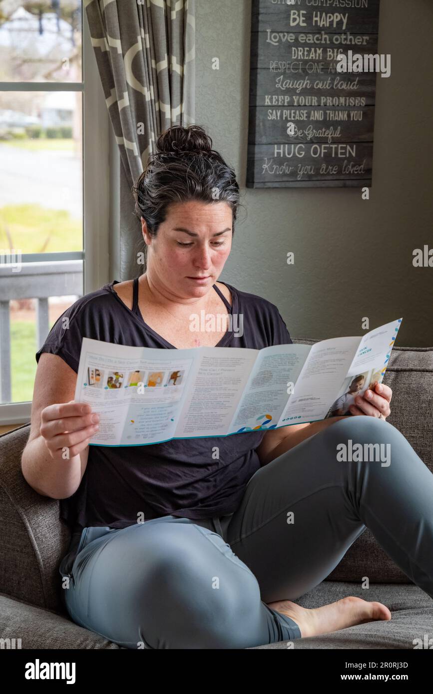 Young 30s female reads skin cancer literature from dermatology department on couch at home upon recent skin cancer diagnosis on her chest. Stock Photo