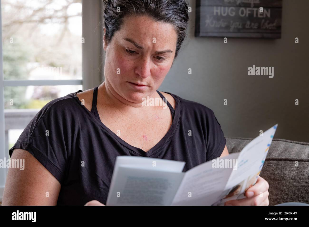 Close-up horizontal image of young 30s female reading skin cancer literature on couch at home upon recent skin cancer diagnosis. Stock Photo