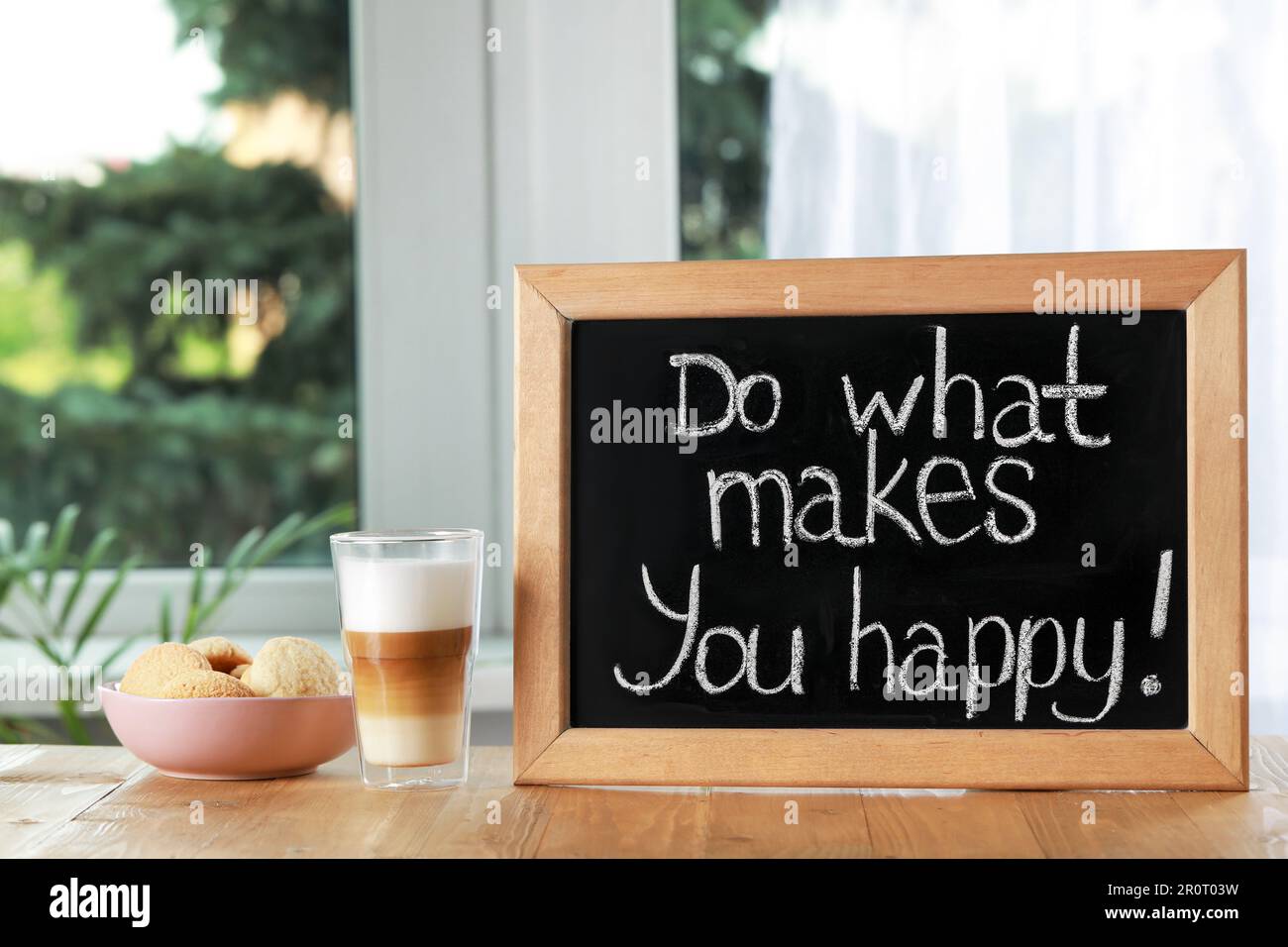 Blackboard with phrase Do What Makes You Happy, coffee and cookies on wooden table near window Stock Photo