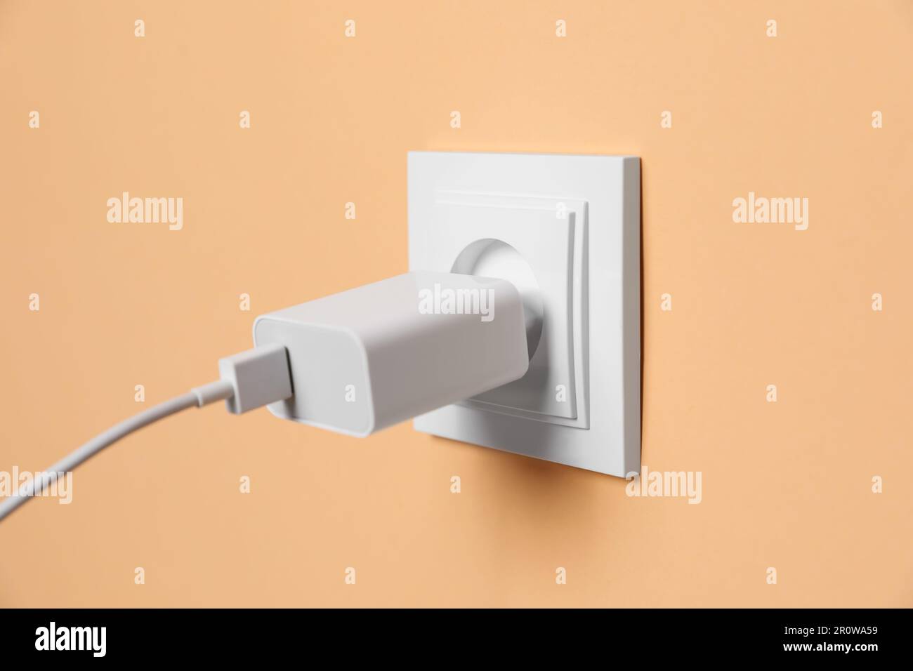 Charger adapter plugged into power socket on pale orange wall, closeup. Electrical supply Stock Photo