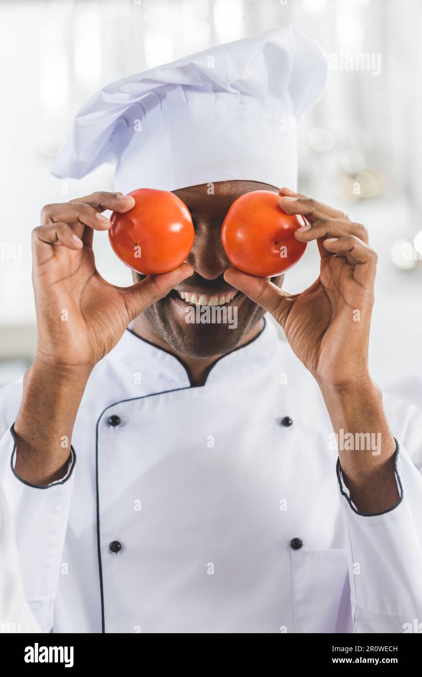 Handsome african american chef covering eyes with tomatoes at restaurant kitchen Stock Photo