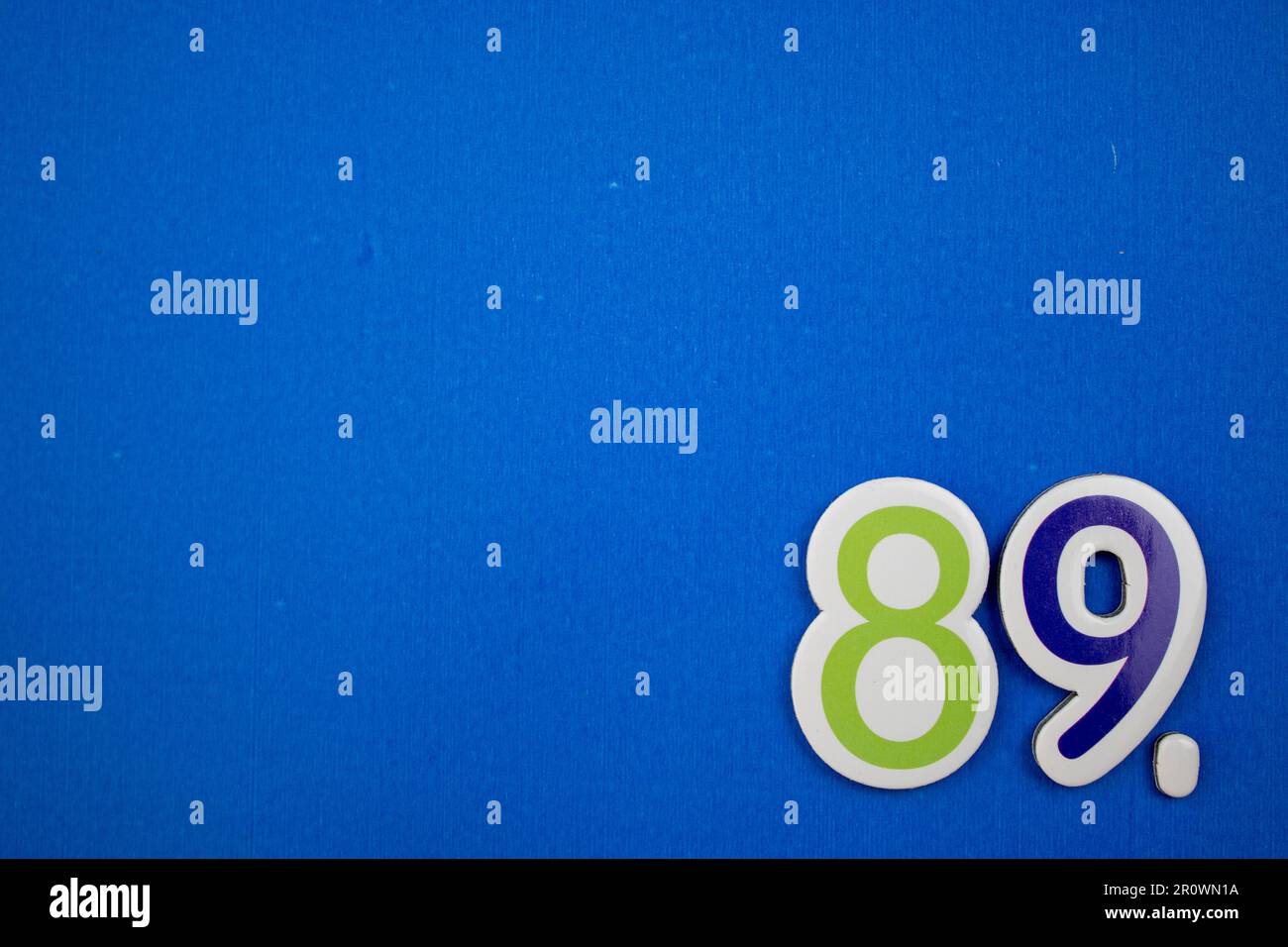 The number 89 in dark blue and green color, photographed from above, placed on the edge of a blue background. Stock Photo