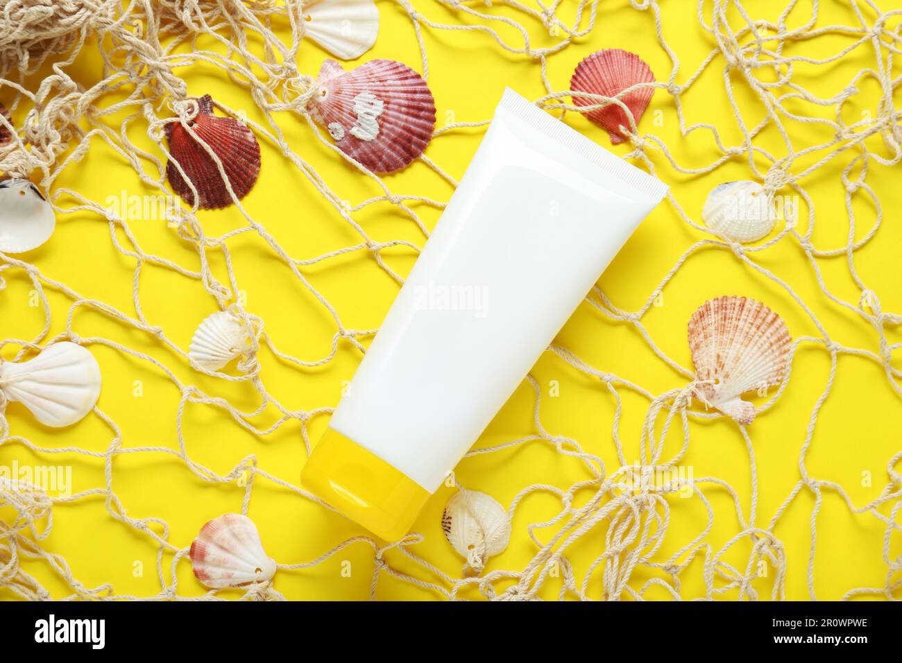 Tube of suntan cream, seashells and net on yellow background, flat lay