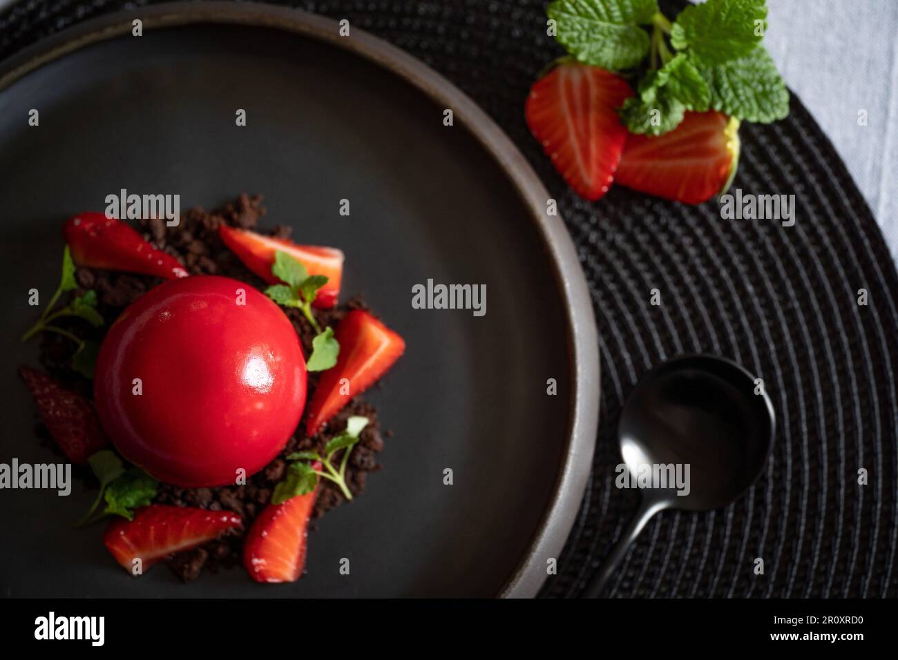 Selective focus of a sphere curd cake with strawberries and brownie. Top view. Dessert with smooth surfaces and mirror glaze. Black plate and spoon. Stock Photo