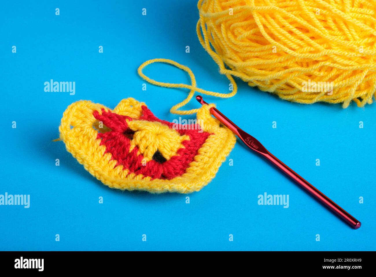 Crochet Granny Square in Yellow and Red with Yellow ball of Yarn and Dark Red Crochet Hook on a Kingfisher blue background. Stock Photo