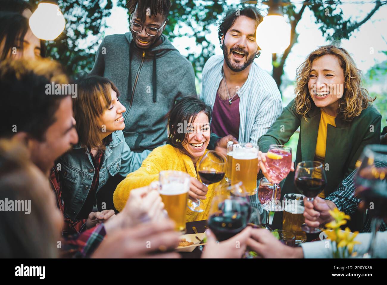 A happy brunette woman holding a glass of red wine is surrounded by her diverse friends, who are raising their own glasses of beer, wine, or cocktails Stock Photo