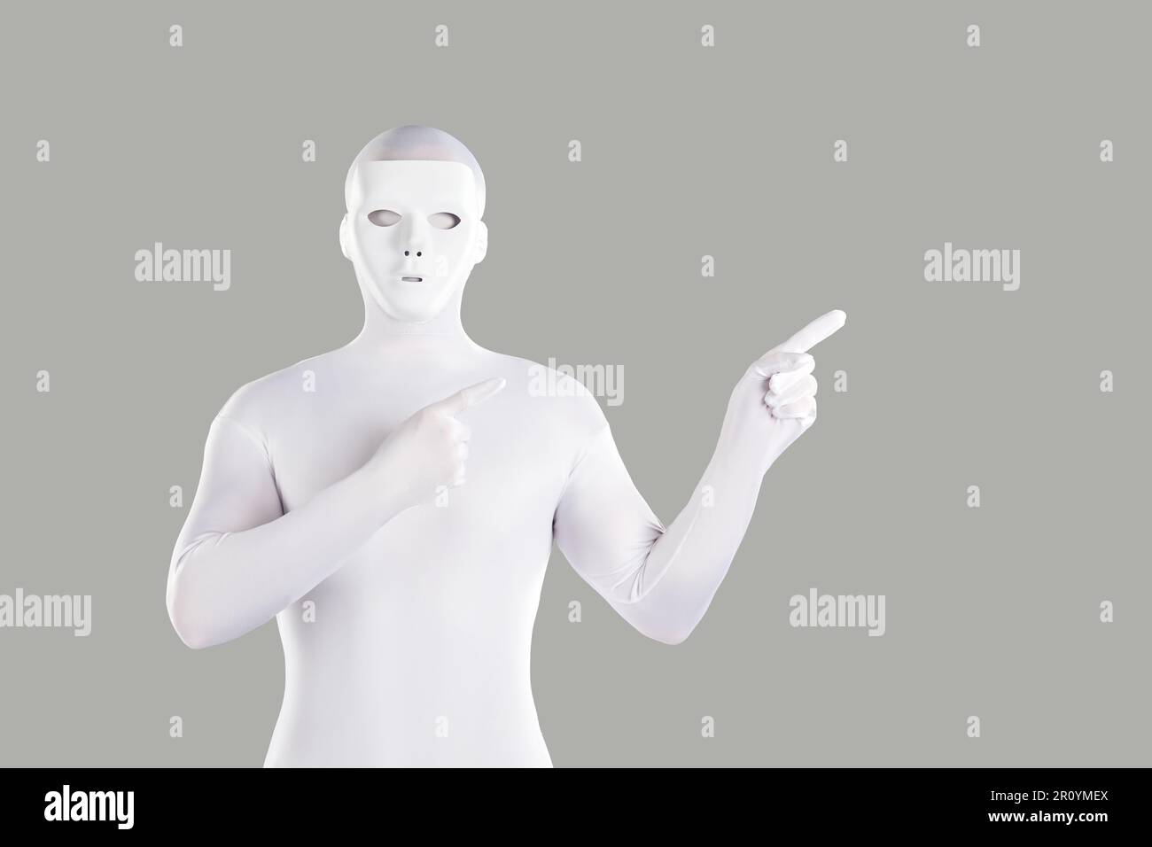 Mysterious man in white mask and spandex bodysuit pointing to side on grey background Stock Photo