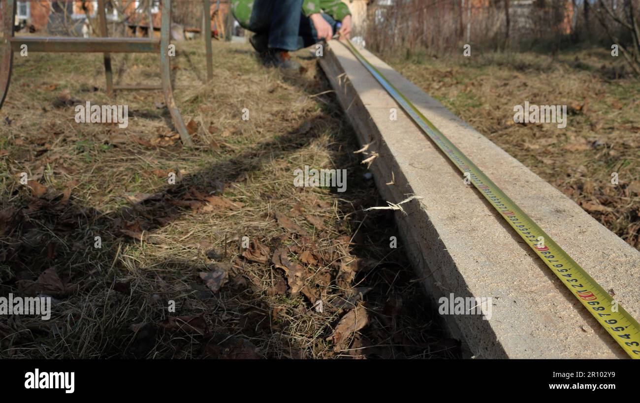 measuring a long board lying on the ground with dry grass using a tape