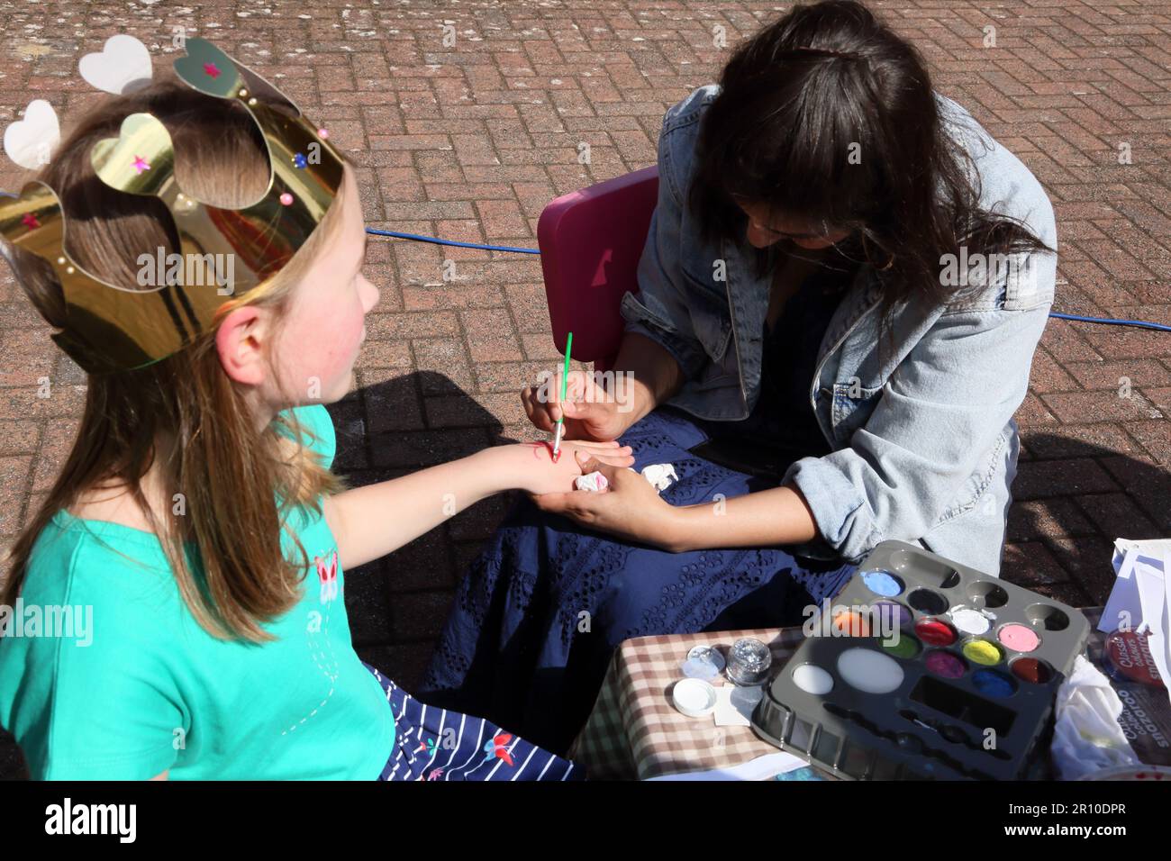 Young Girl Wearing Paper Crown with Woman Painting on her hand with Face Paints at Street Party Celebrating King Charles III Coronation Surrey England Stock Photo
