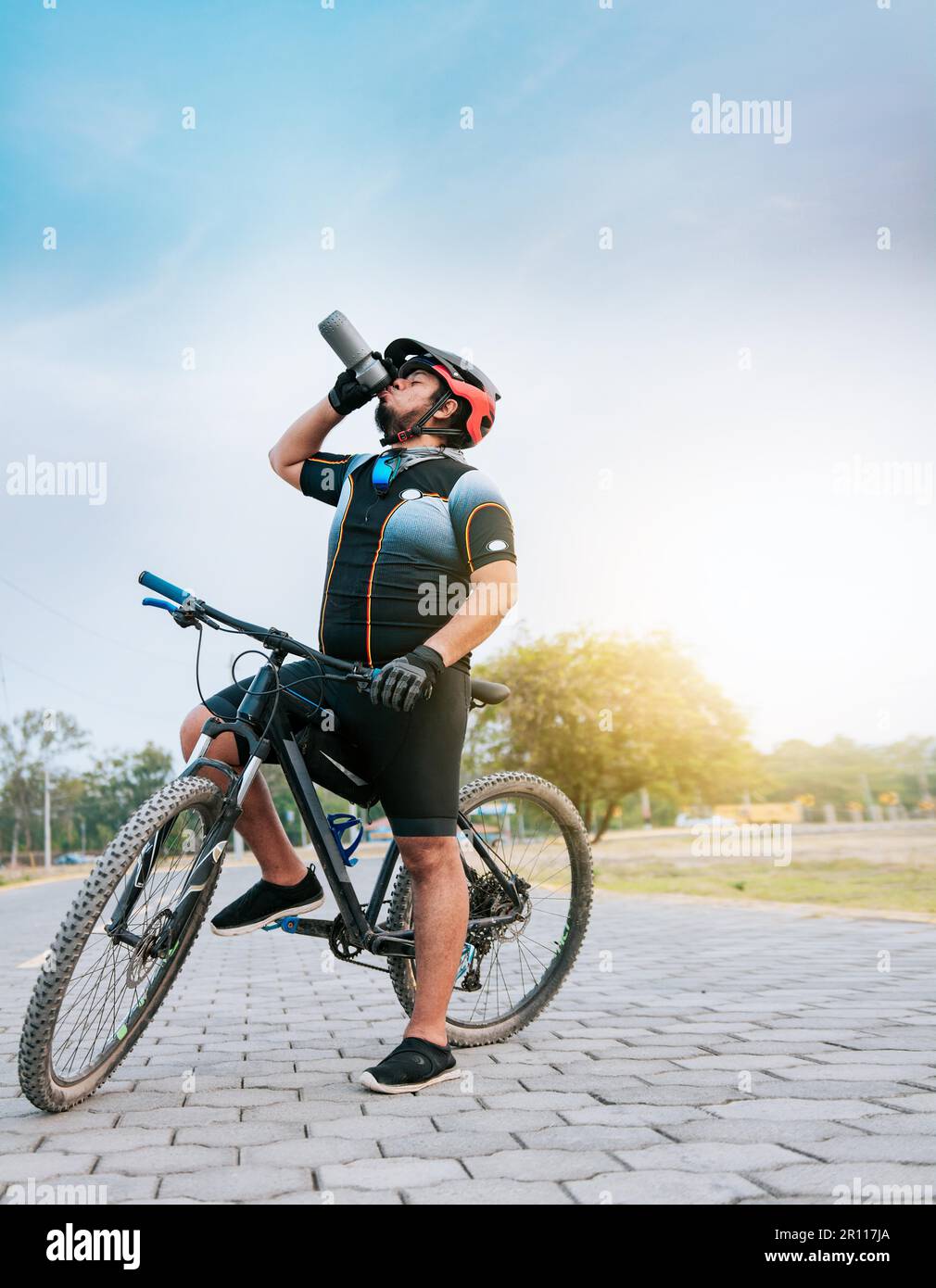 Thirsty cyclist on his bike drinking water outside. Tired cyclist drinking water outdoors, Chubby cyclist on his bike drinking water Stock Photo