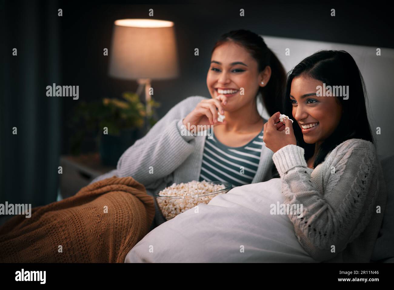 Laughing, popcorn and women watching tv, movies or show at home for sleepover with streaming service and comedy. Gen z people relax in bedroom with Stock Photo