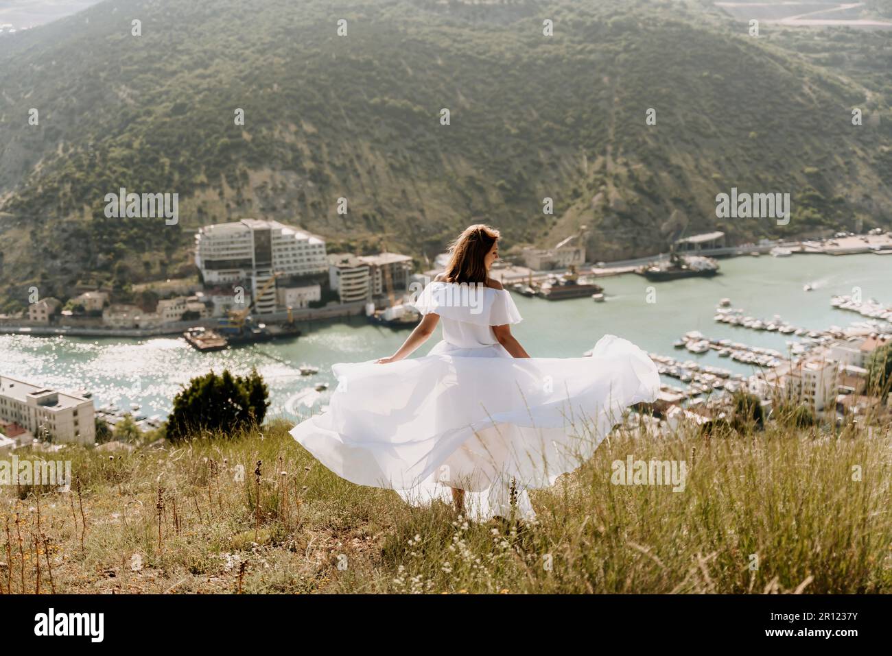 Happy woman in a white dress and hat stands on a rocky cliff above the ...
