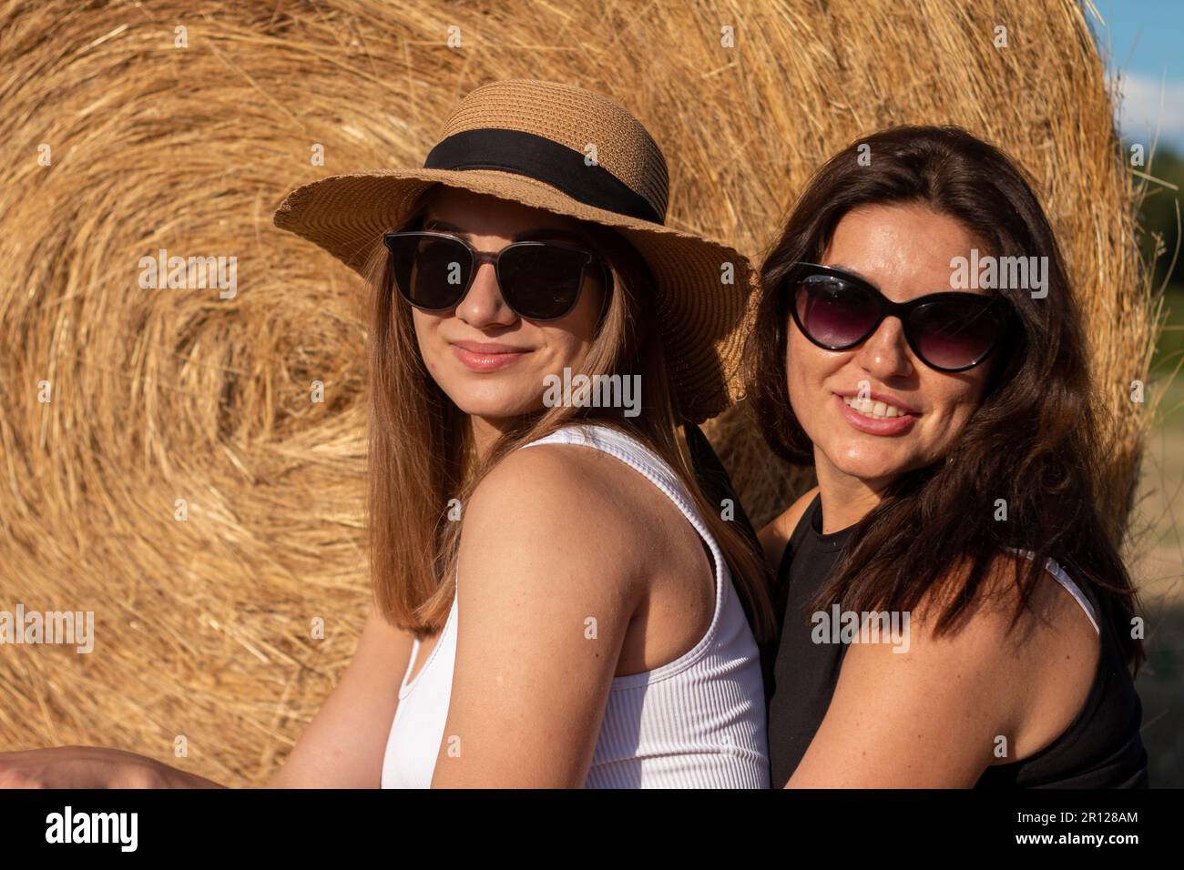 Two young women smiling and enjoying the day wearing stylish wide-brimmed hats and sunglasses while sitting side by side Stock Photo