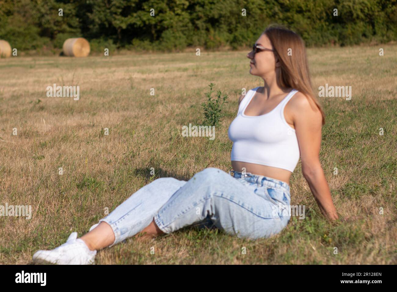 A young woman with blonde hair enjoying a sunny day outdoors, seated on a grassy field in a white tank top and blue jeans Stock Photo