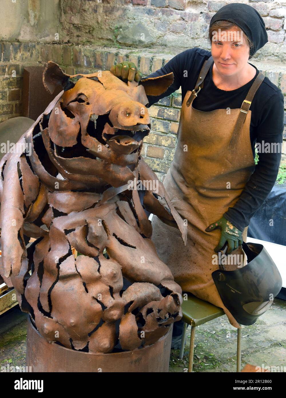 PRODUCTION - 05 May 2023, Saxony, Leipzig: Steel artist Dana Meyer stands in her backyard, which is part of the studio, at a 1.30-meter-high pig of an installation with six large and small domestic pigs. Along with smaller metal insects in glass, the unusual life-size steel sculptures are among the works that the artist forges freehand and without plastic designs. She uses mostly found steel plates to create forged parts, which she assembles into the unusual figures like a puzzle using a cutting torch and welding equipment. She is represented with her works from 12.5.-20.08.23 at the Galerie R Stock Photo