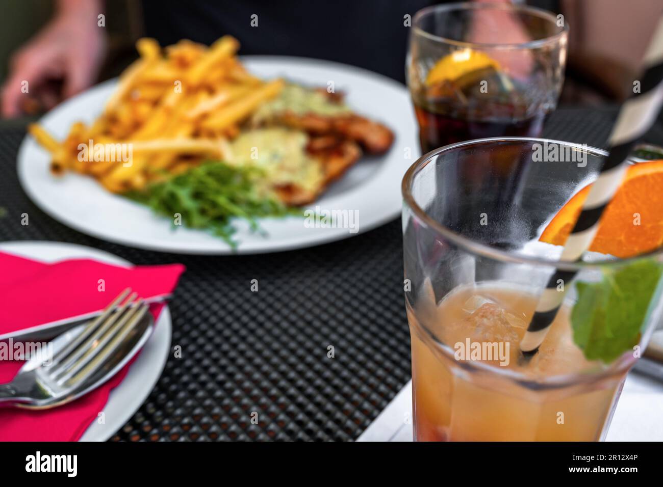 Table with fruity drink and lemonade, plate with cutlery and plate with food (french fries, meat, sauce and rucola leaf). Dining in outdoor restaurant Stock Photo