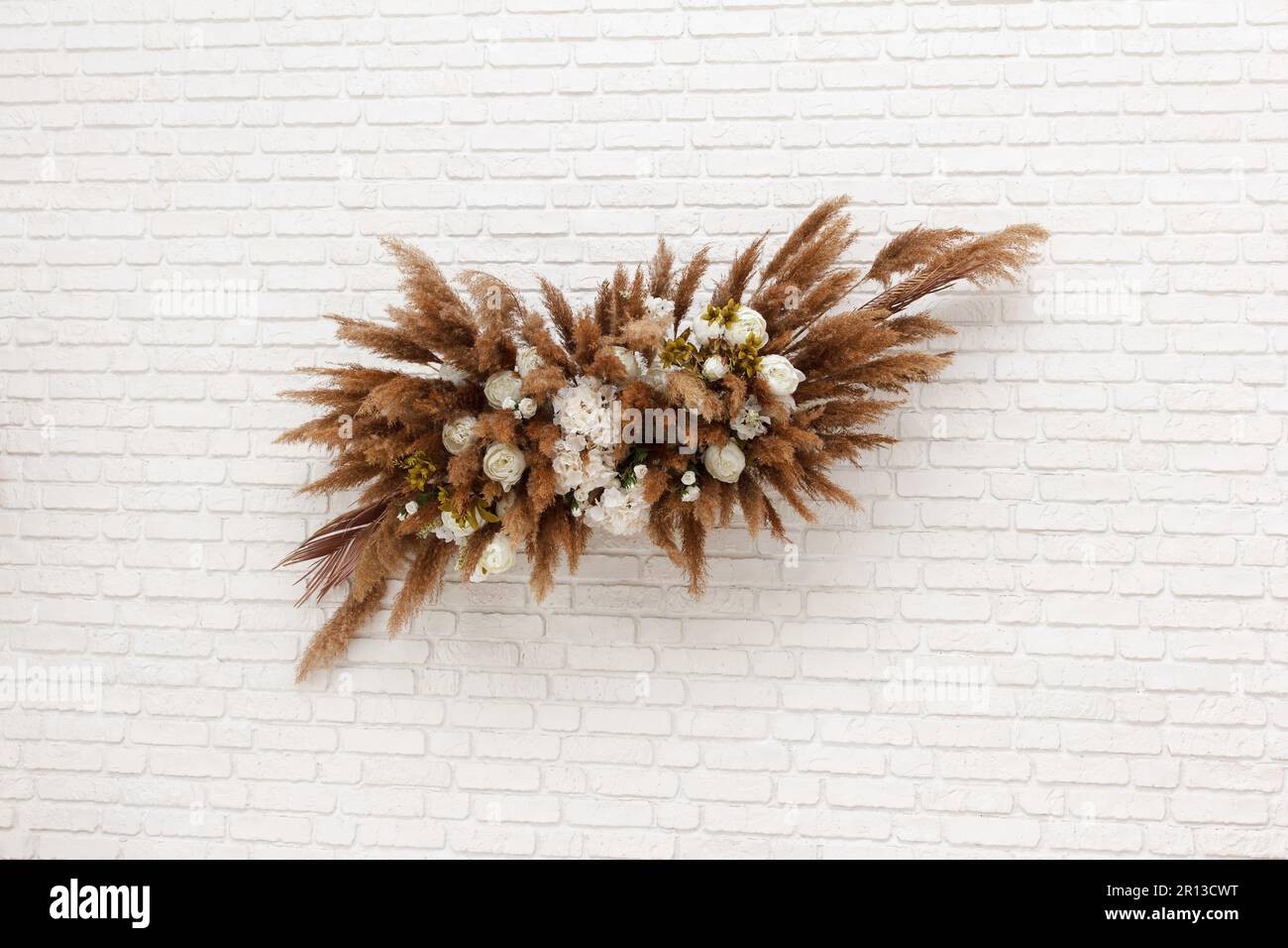 A beautiful floral set of white flowers, reeds and feathers in white and brown colors hangs on a white brick wall. And beautifully adorns interior of Stock Photo