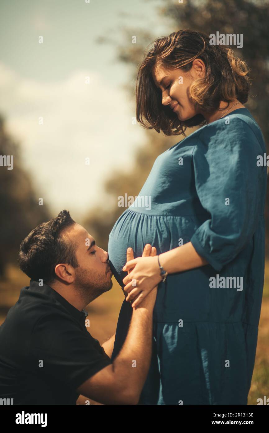 A tender moment as a man in a black t-shirt affectionately kisses his pregnant wife's belly, both standing in an olive field Stock Photo