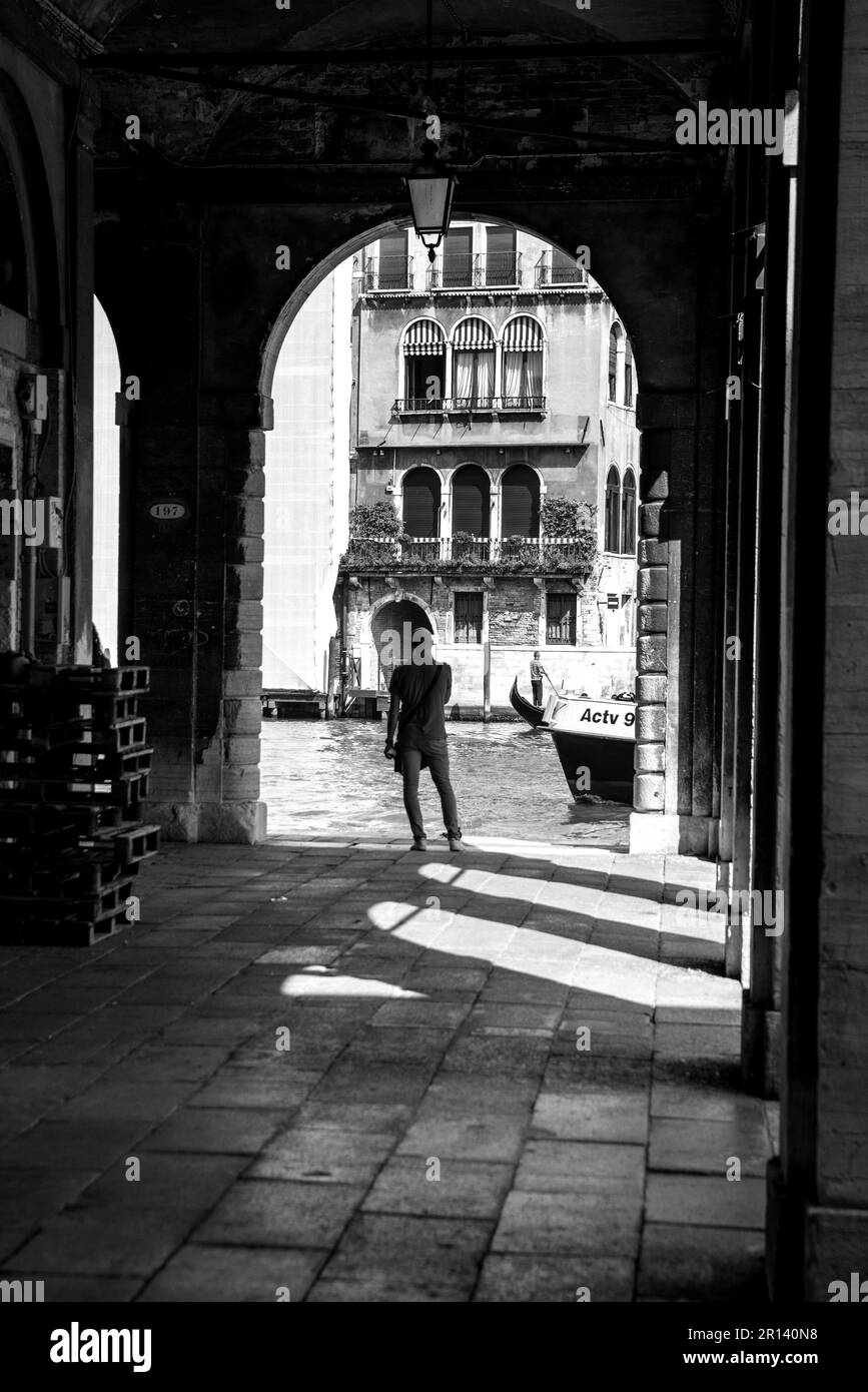 A lone man in the shadow of stone arches looking over a canal in Venice, Italy (Black and white) Stock Photo