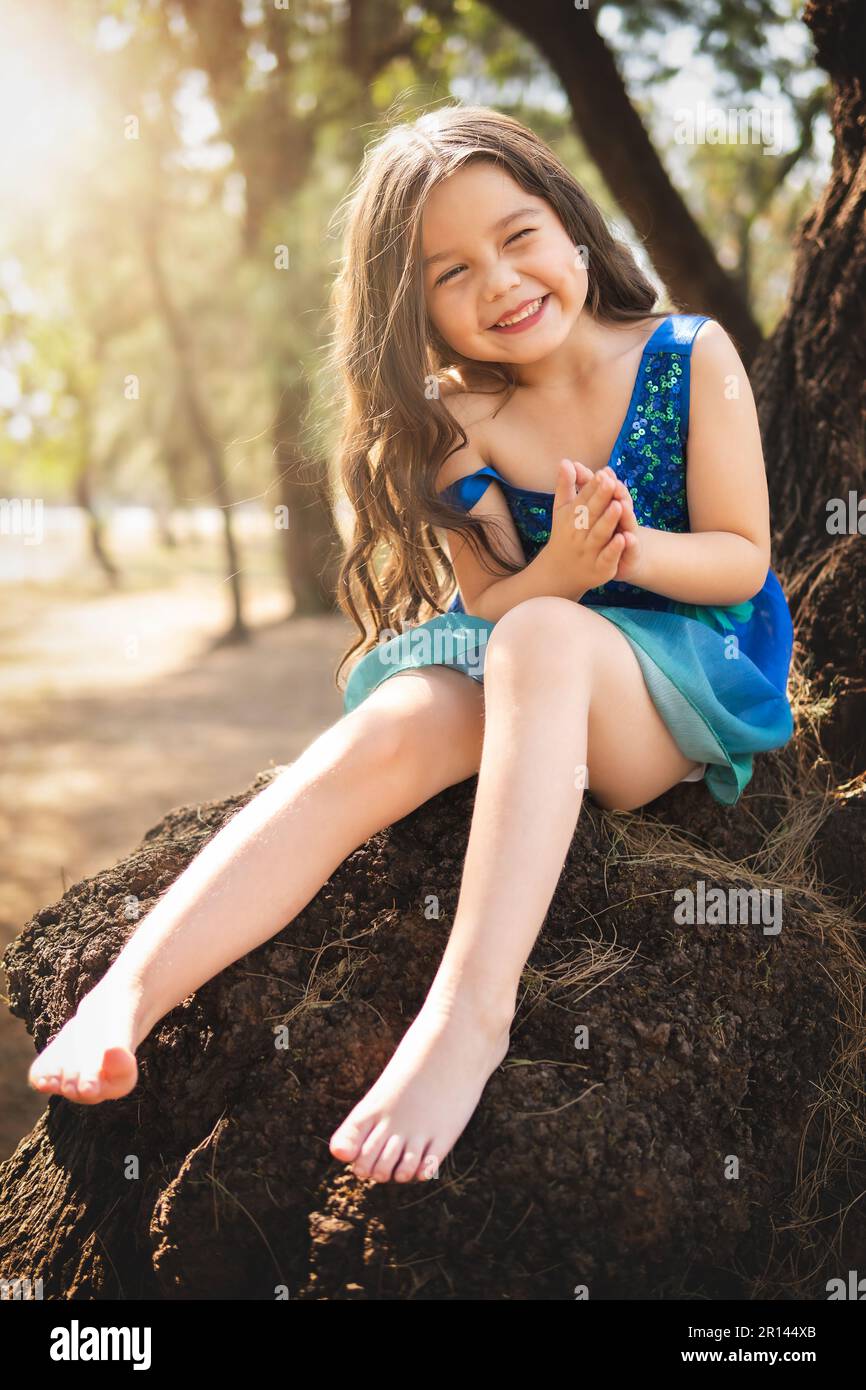 beautiful happy girl playing in the park with blue dress without shoes sitting on tree laughing screaming with happiness in family enjoying children's Stock Photo