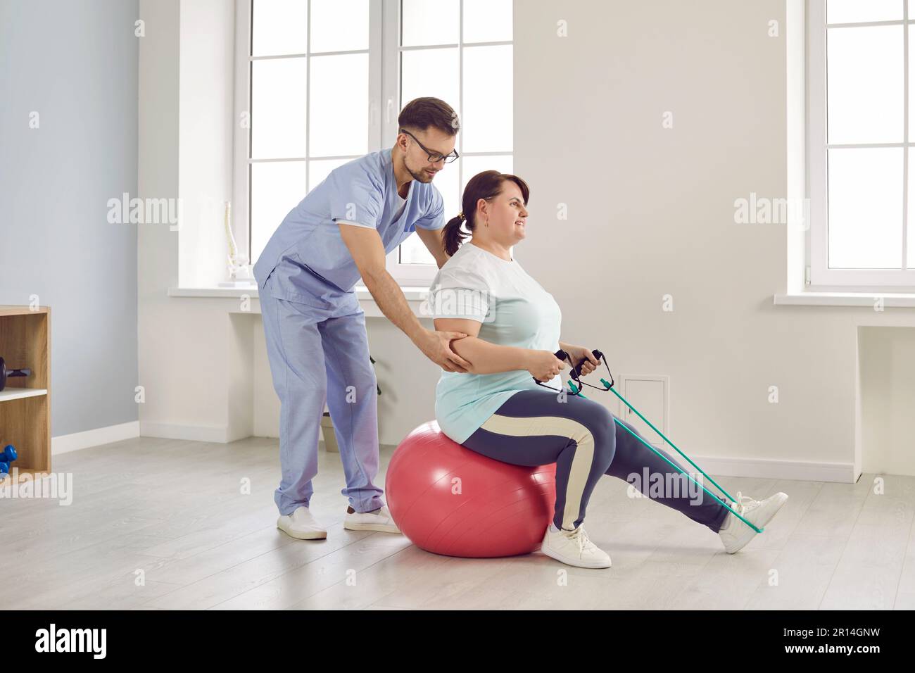 Nurse man helping fat patient woman to do stretching exercises with rubber band sitting on fit ball Stock Photo