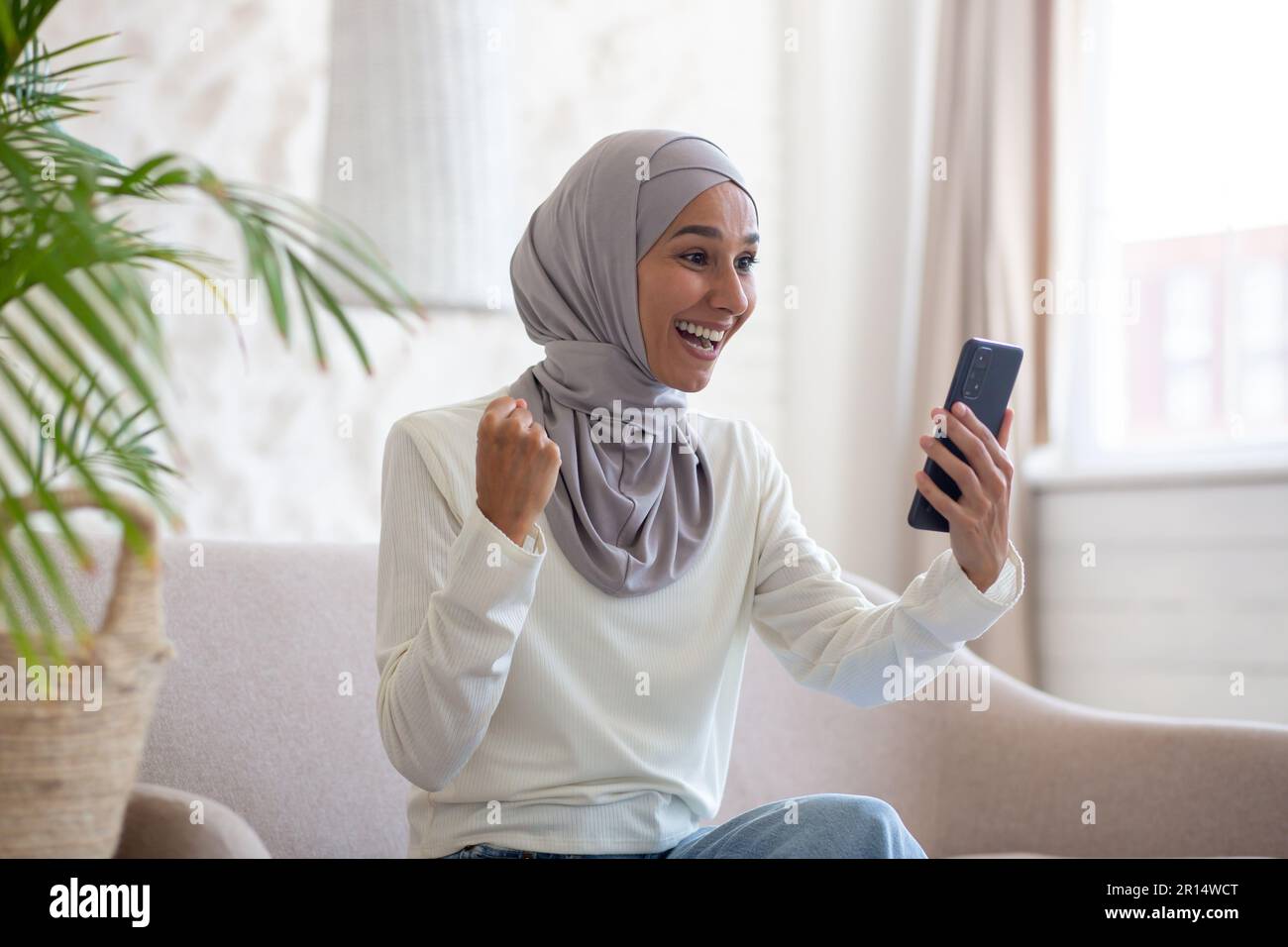 Happy young Arab woman in hijab sitting on sofa at home and using ...