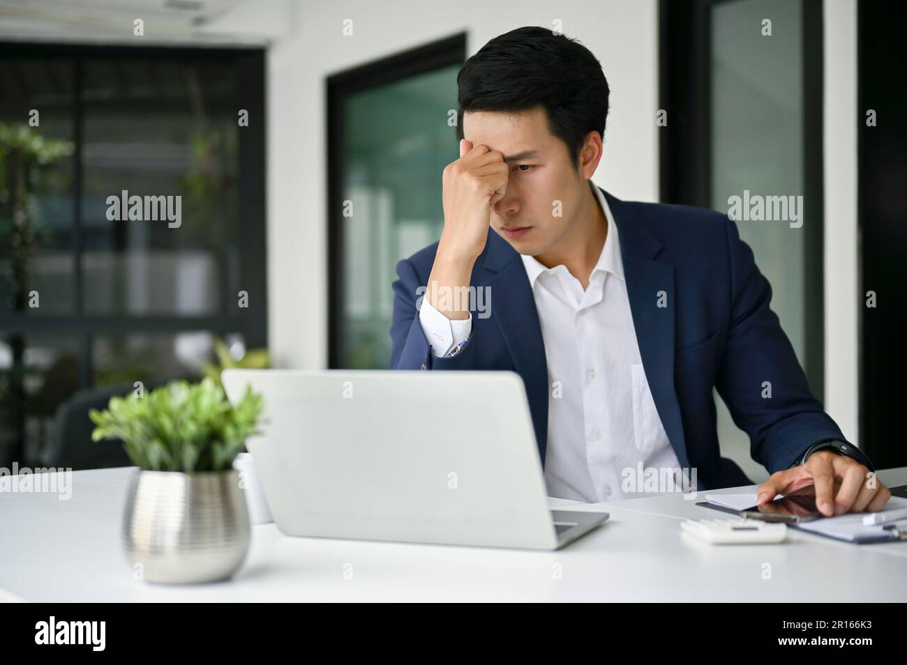 Stressed millennial Asian businessman in formal business suit sits at his desk, looking art his laptop screen and pensive thinking a plan to solve the Stock Photo