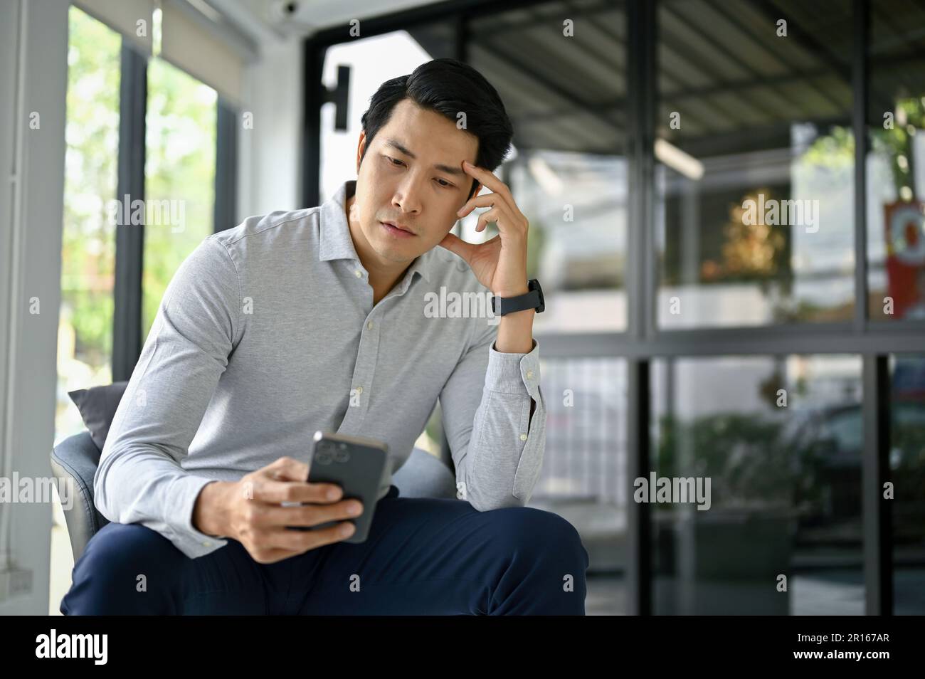 Stressed and thoughtful millennial Asian businessman or male boss reading messages on his smartphone while sitting in his office. Stock Photo