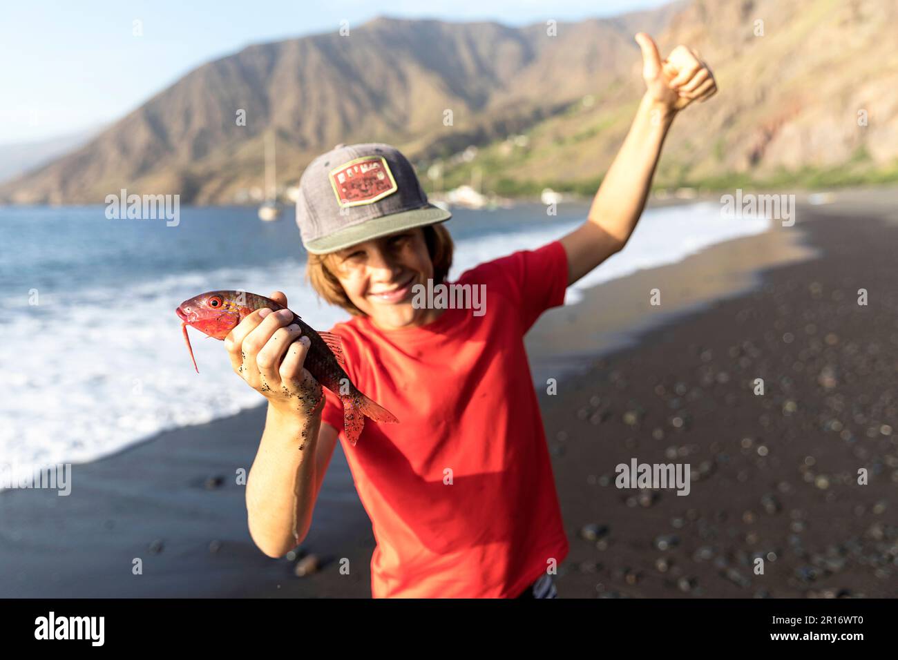 Happy boy holding a red fish that he caught in one hand and thumbs up with the other one on black sandy beach, Tarrafal, Santo Antao, Cabo Verde Stock Photo
