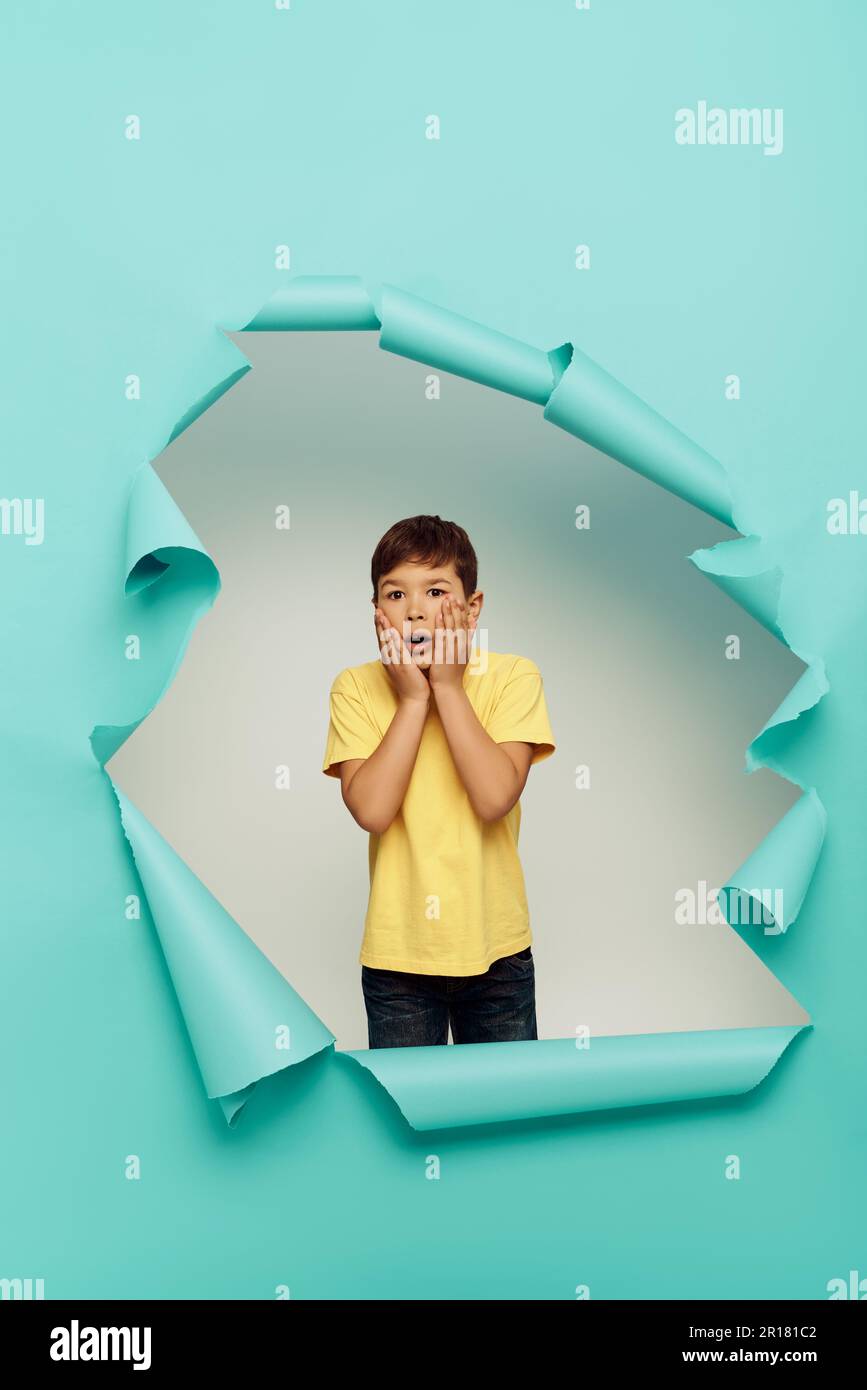 Shocked multiracial boy in yellow t-shirt touching cheeks and looking at camera during world child protection day while standing behind hole in blue p Stock Photo