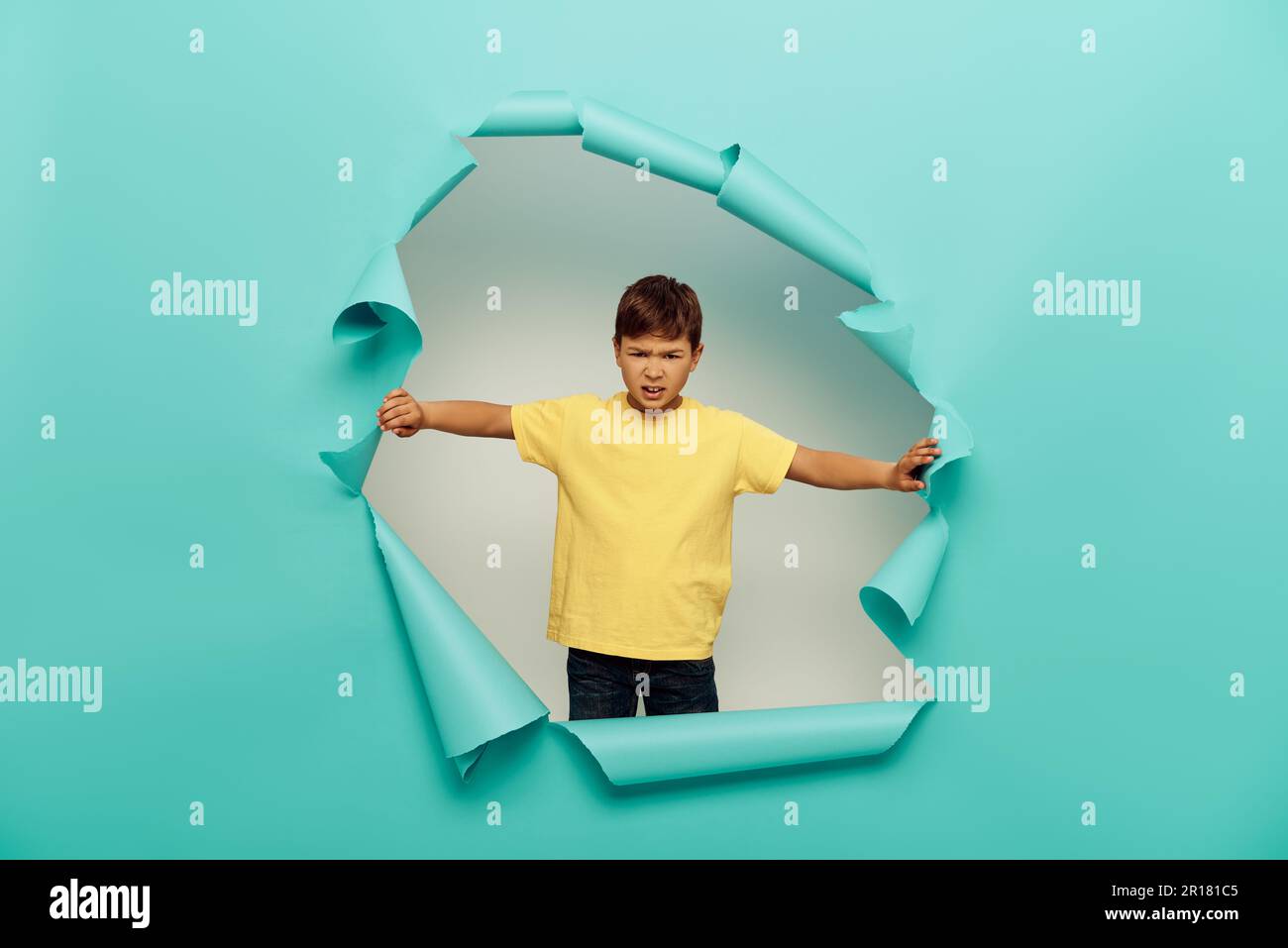 Angry multiracial boy in yellow t-shirt looking at camera during international child protection day while standing behind hole in blue paper on white Stock Photo