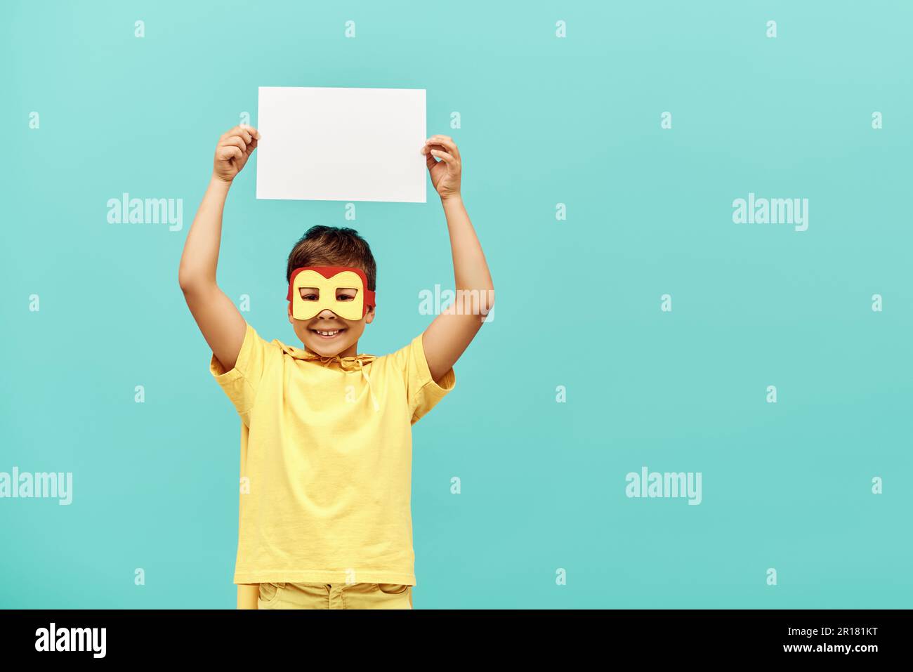 smiling multiracial boy in yellow superhero costume with mask holding blank paper above head on blue background, International children's day concept Stock Photo