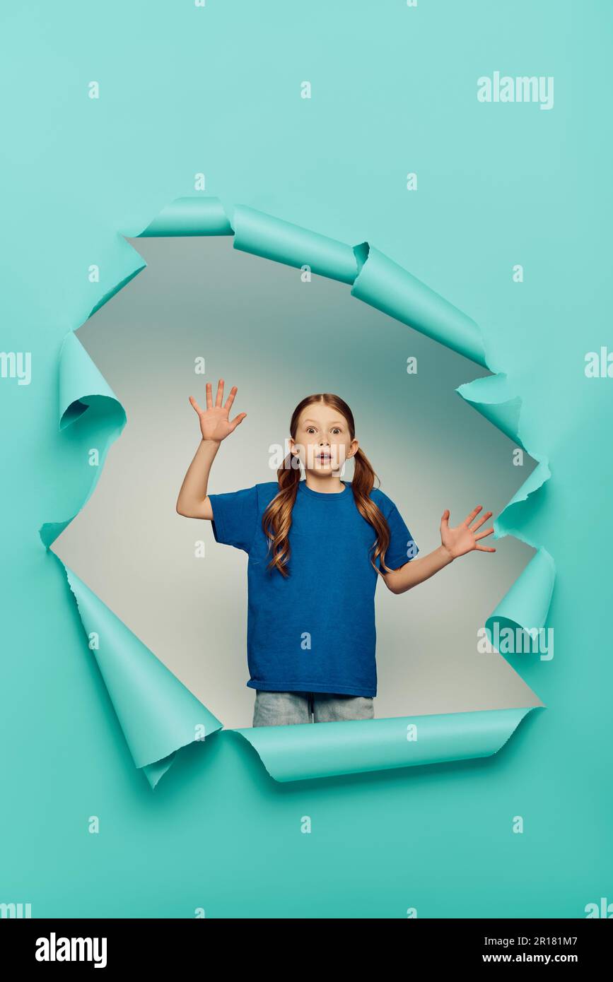 terrified redhead preteen girl looking at camera while gesturing near blue torn paper hole on white background, International children's day concept Stock Photo
