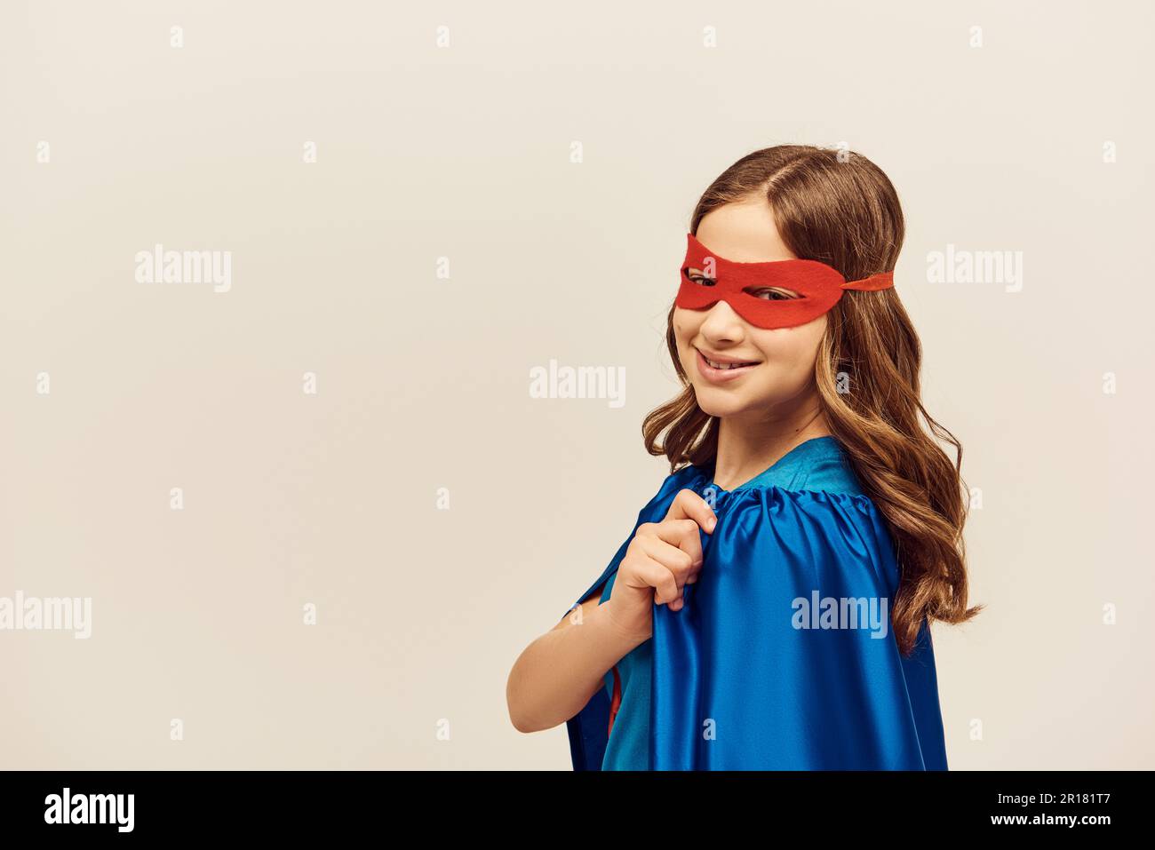 happy girl in superhero costume with blue cloak and red mask on face looking at camera and smiling while celebrating International children's day on g Stock Photo