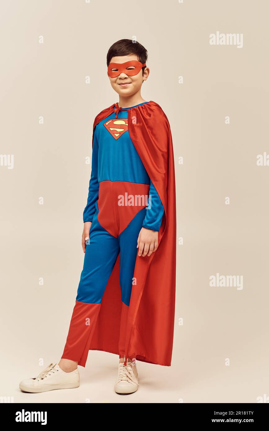 full length of happy asian boy in red and blue superhero costume with cloak and mask on face smiling while celebrating International children's day ho Stock Photo