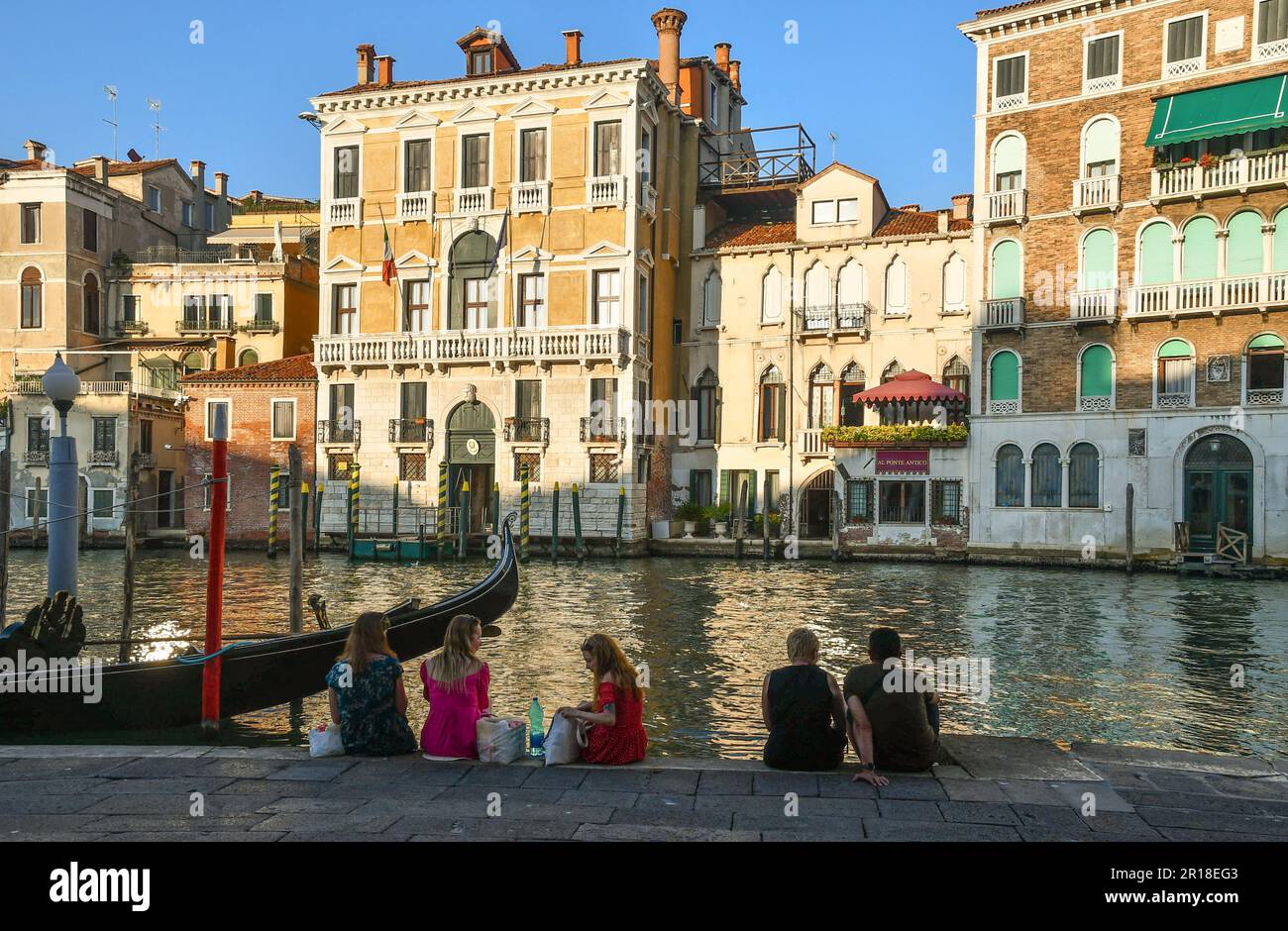 Three friends and a couple resting on the shore of the Grand Canal next to a a moored gondola in summer, Venice, Veneto, Italy Stock Photo