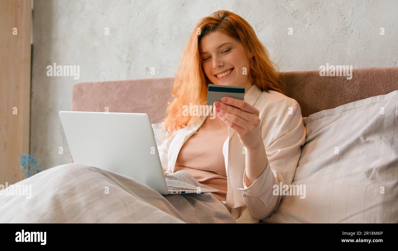 Happy female cardholder with credit card bank payment with laptop lying in bed at morning woman consumer buyer girl paying for purchase in web store Stock Photo