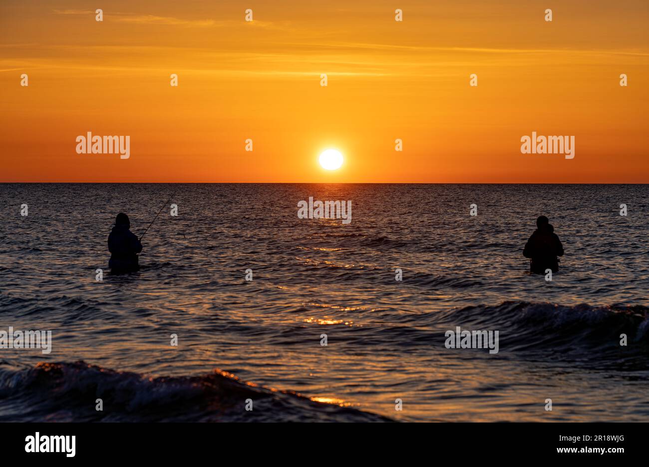 The silhouettes of two anglers in waterproof trousers fishing in front of a dreamy orange sunset in the Baltic Sea. They are standing in the middle of Stock Photo