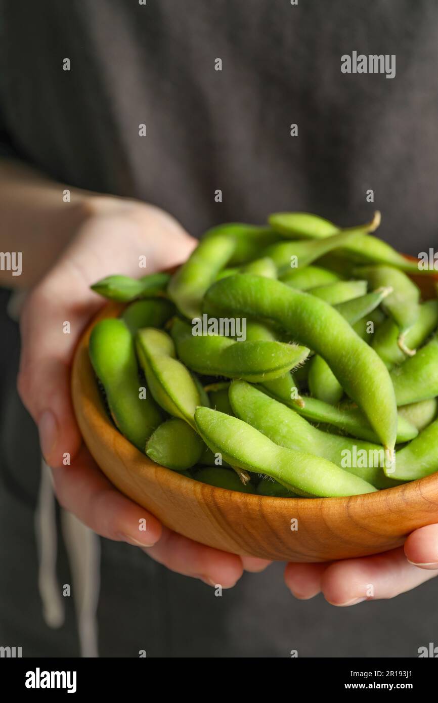 Woman holding bowl with green edamame beans in pods on black background