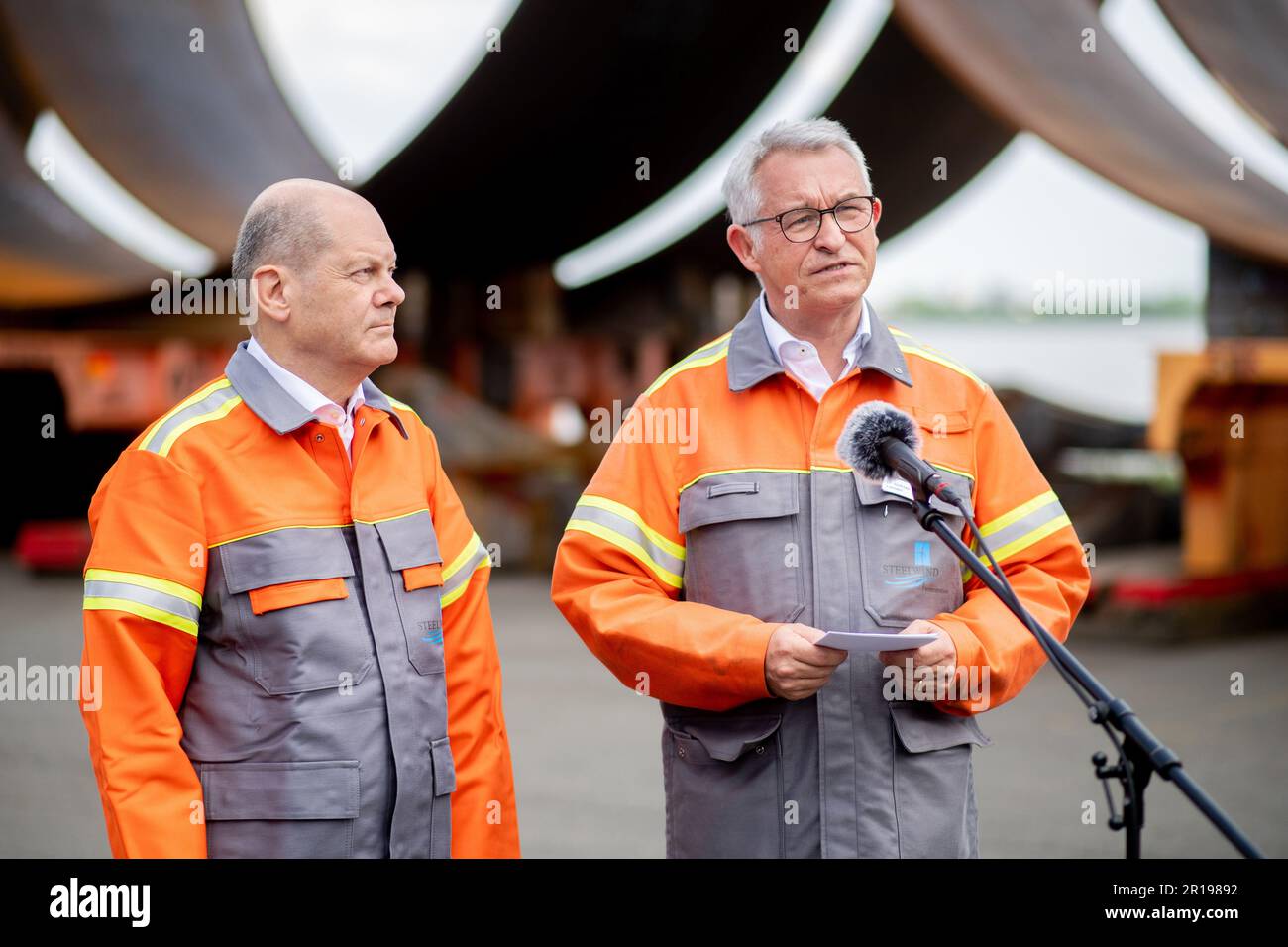 Nordenham, Germany. 12th May, 2023. Andreas Liessem (r), Managing Director of Steelwind Nordenham GmbH, speaks to journalists while standing with German Chancellor Olaf Scholz (SPD) in front of numerous components for the steel foundations of wind turbines. Scholz visited the production of steel foundations, so-called monopiles, for wind turbines in offshore wind farms at Steelwind Nordenham GmbH. Credit: Hauke-Christian Dittrich/dpa/Alamy Live News Stock Photo