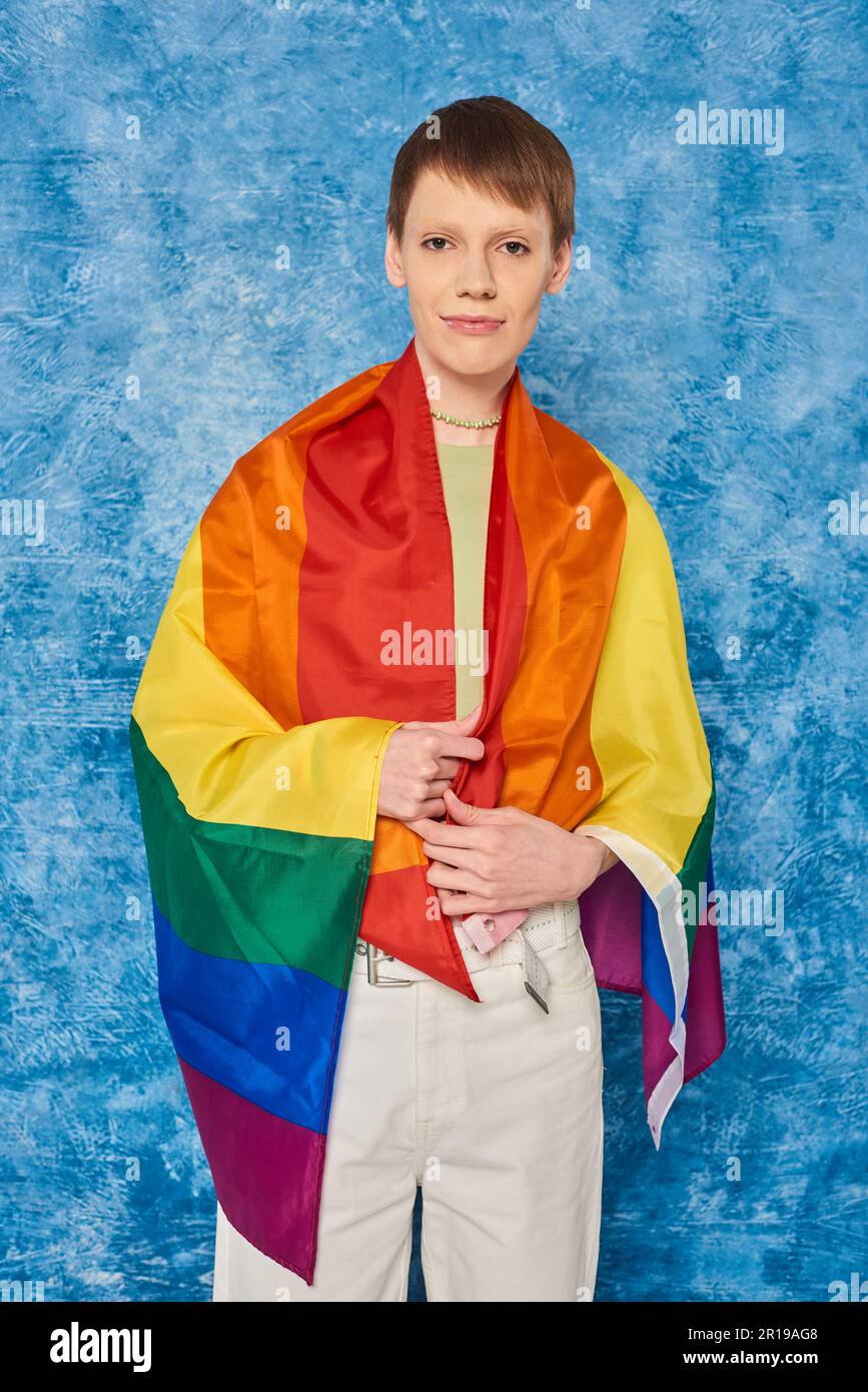 Portrait of young queer person wrapped in lgbt flag looking at camera