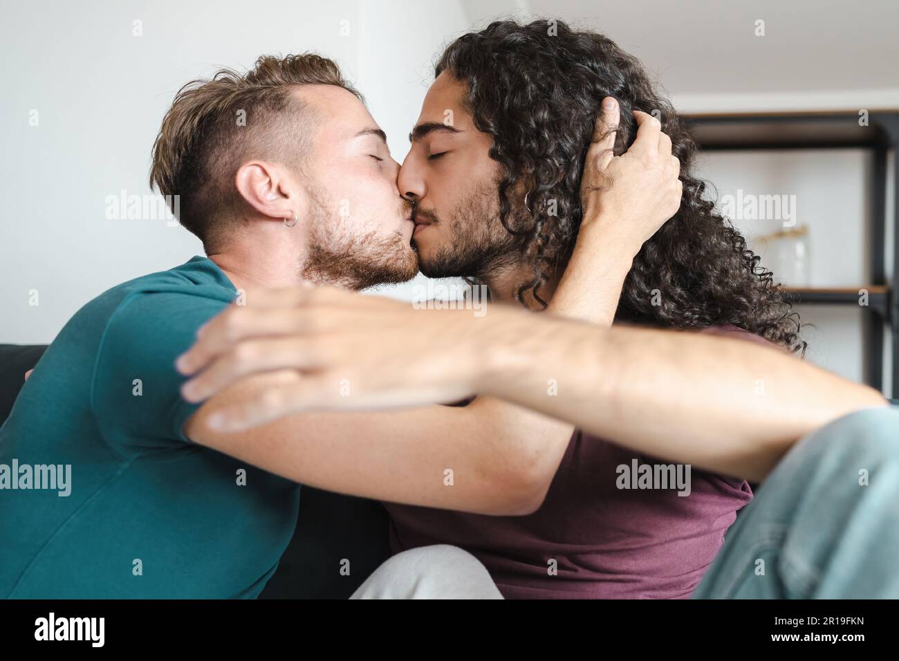 A loving gay couple, one with short light hair and the other with long dark hair, share a tender kiss on the lips while sitting on a sofa, both wearin Stock Photo