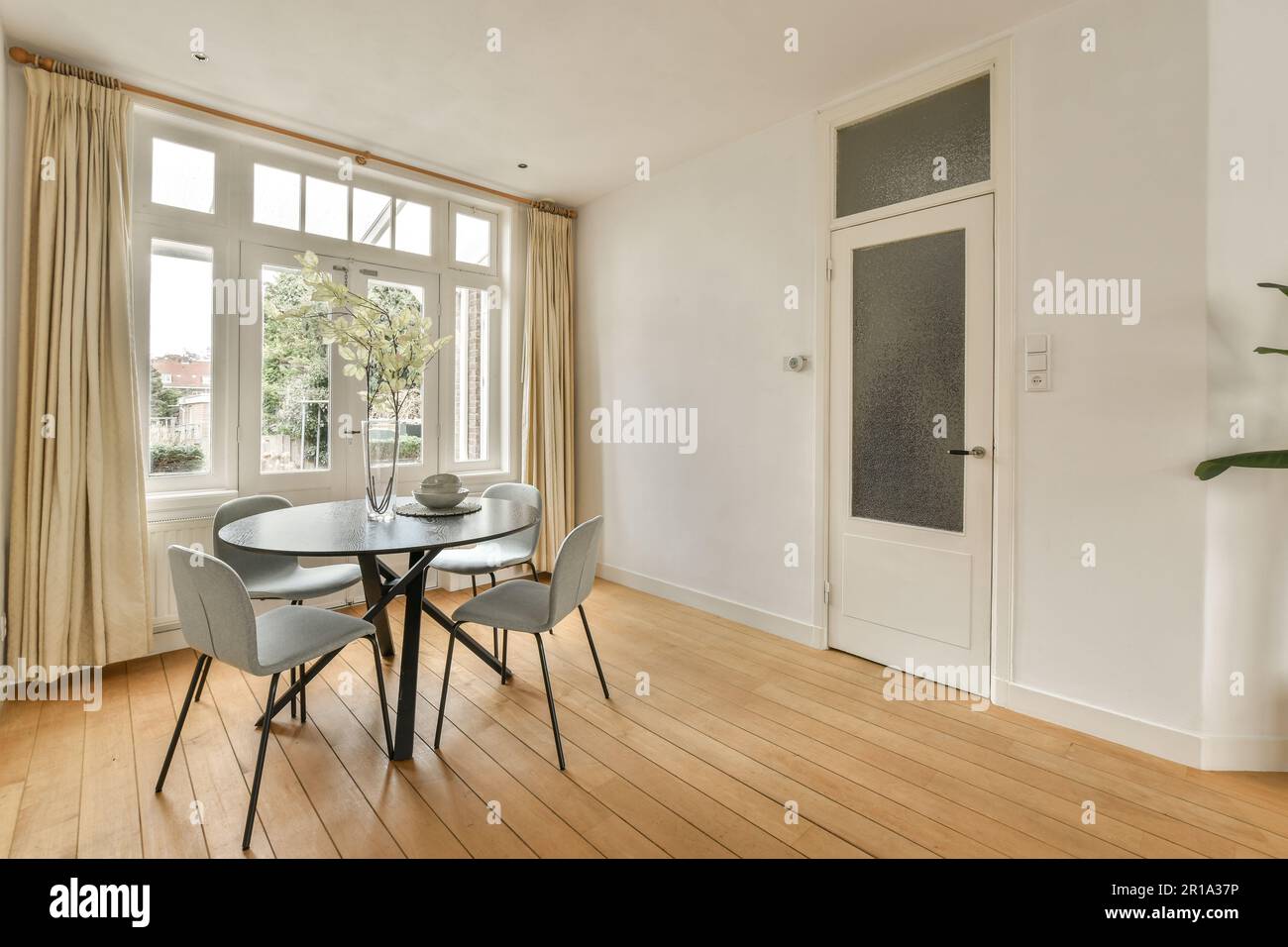 a dining table and chairs in a room with large windows looking out onto the street below, there is a pot of flowers on Stock Photo
