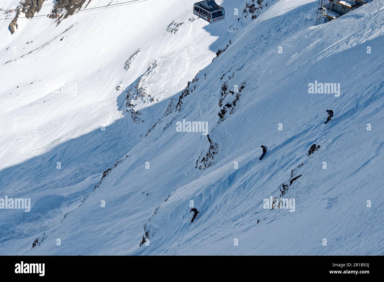Three  Snowboarders start their descent while a cable car is reaching the peak in the swiss alps Stock Photo