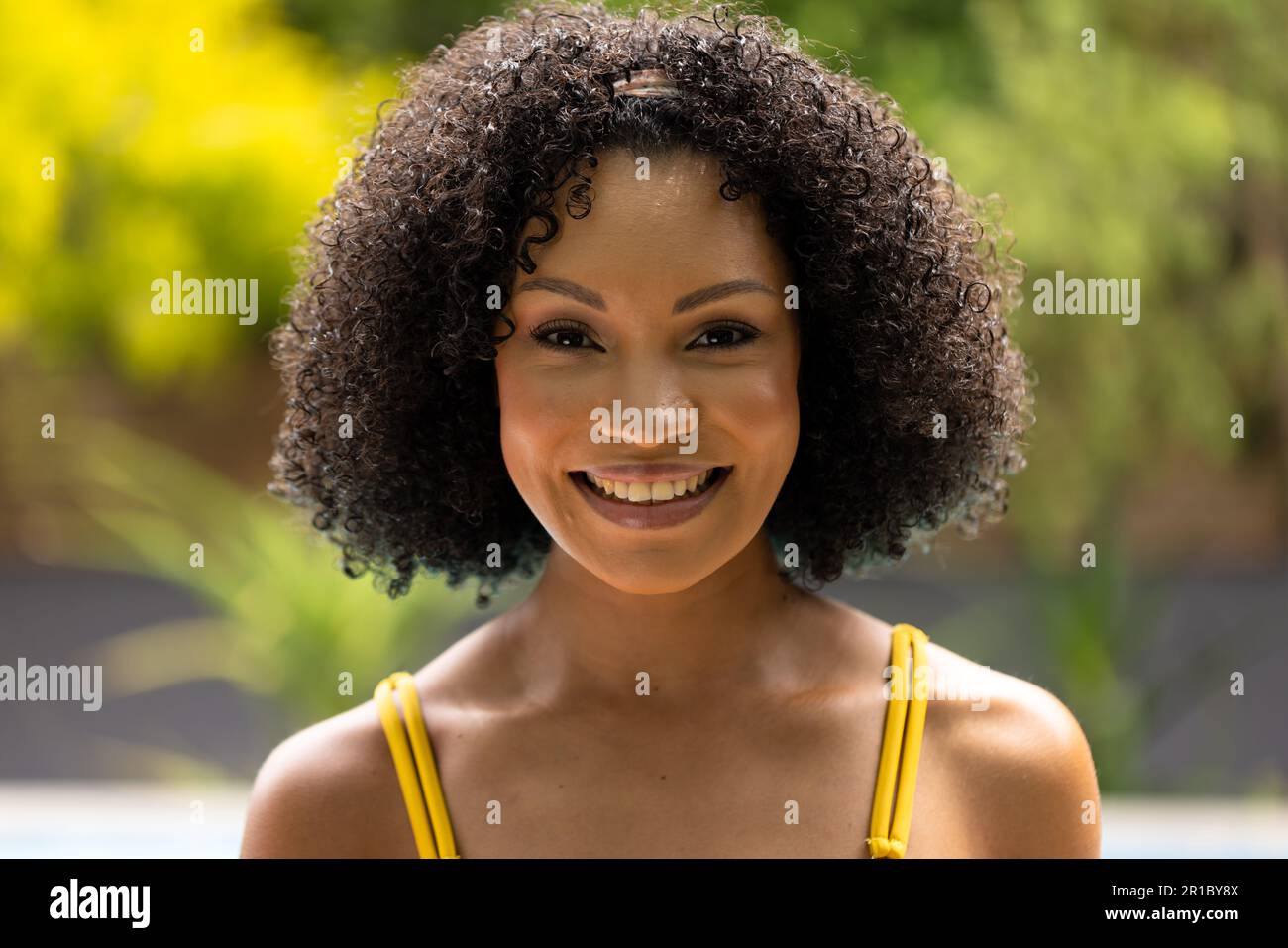 Portrait of smiling african american woman in swimsuit standing by pool in garden Stock Photo